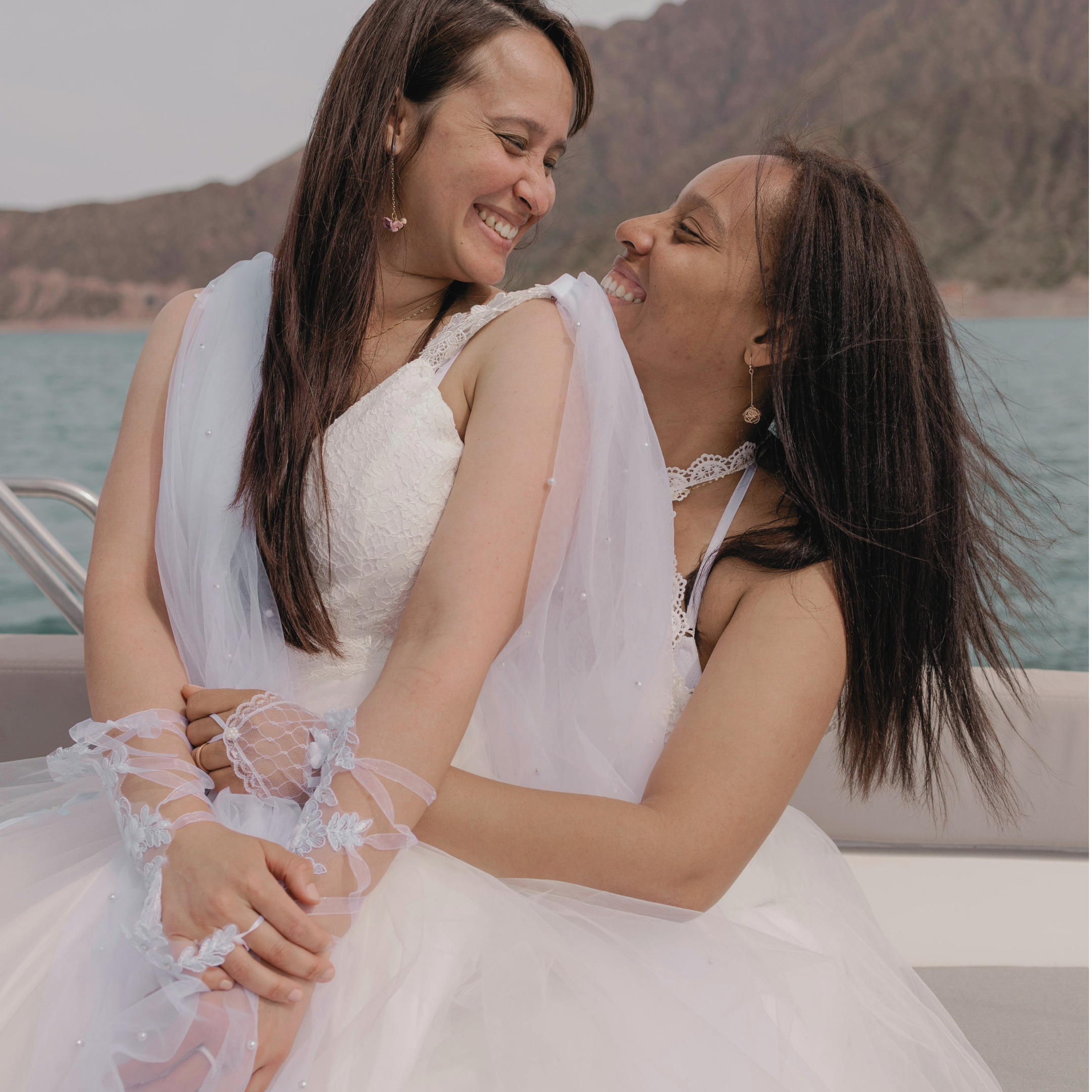 Two women in white dresses smiling and hugging each other outdoors with mountains in the background.