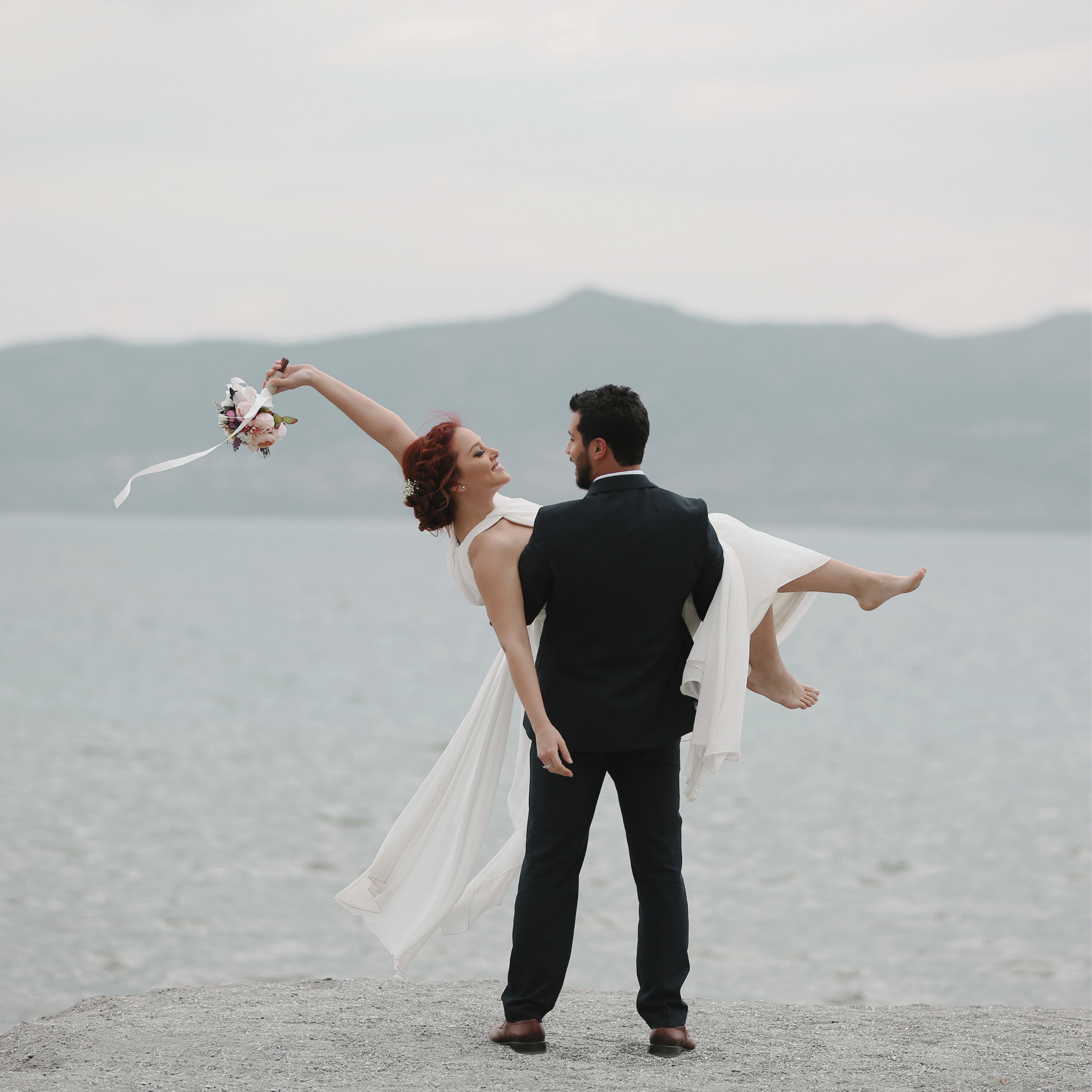 A bride and groom standing on a beach with a mountain range in the background, the bride is holding a bouquet and being lifted by the groom, they are dressed in wedding attire.