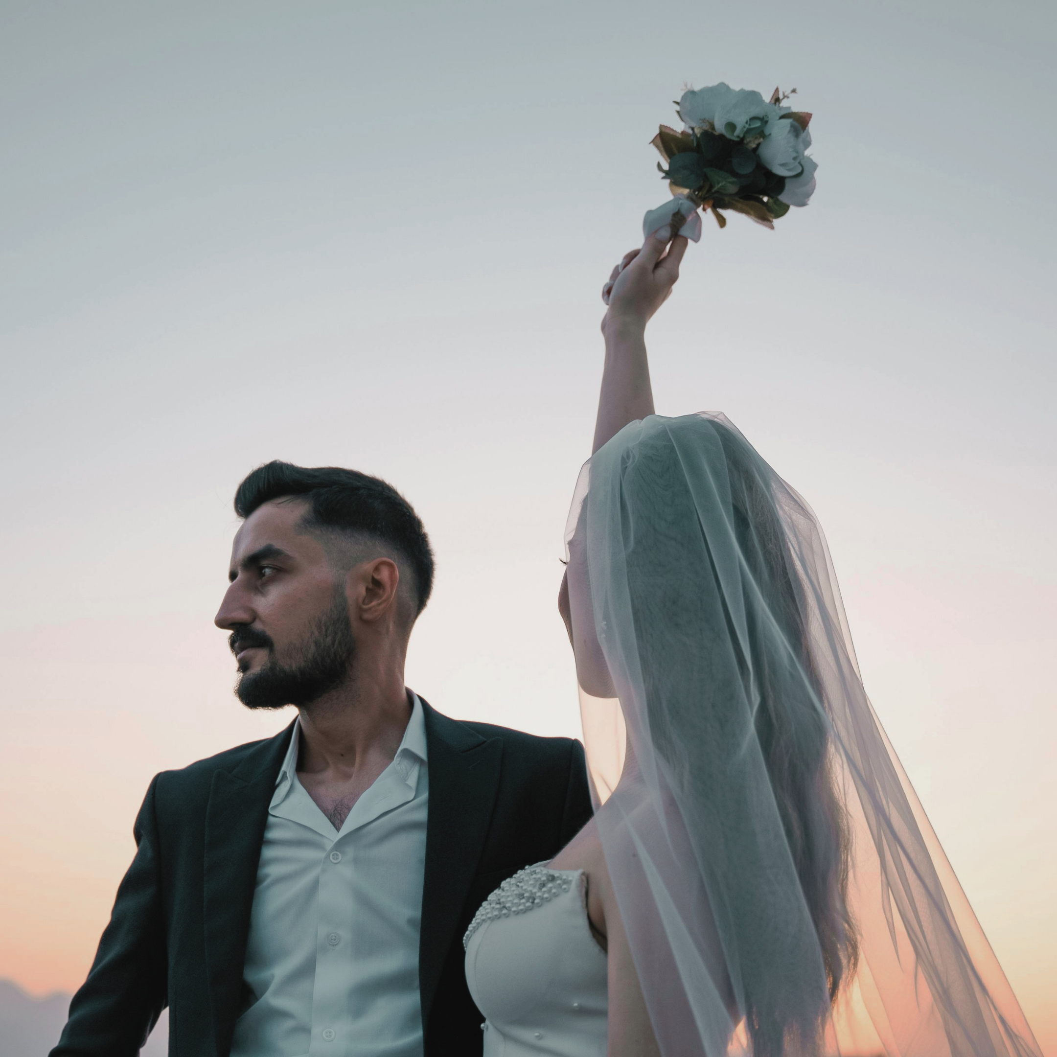A close-up of a couple, with the man in a suit and the woman in a wedding dress, raising a bouquet into the air against a pastel sky at sunset.