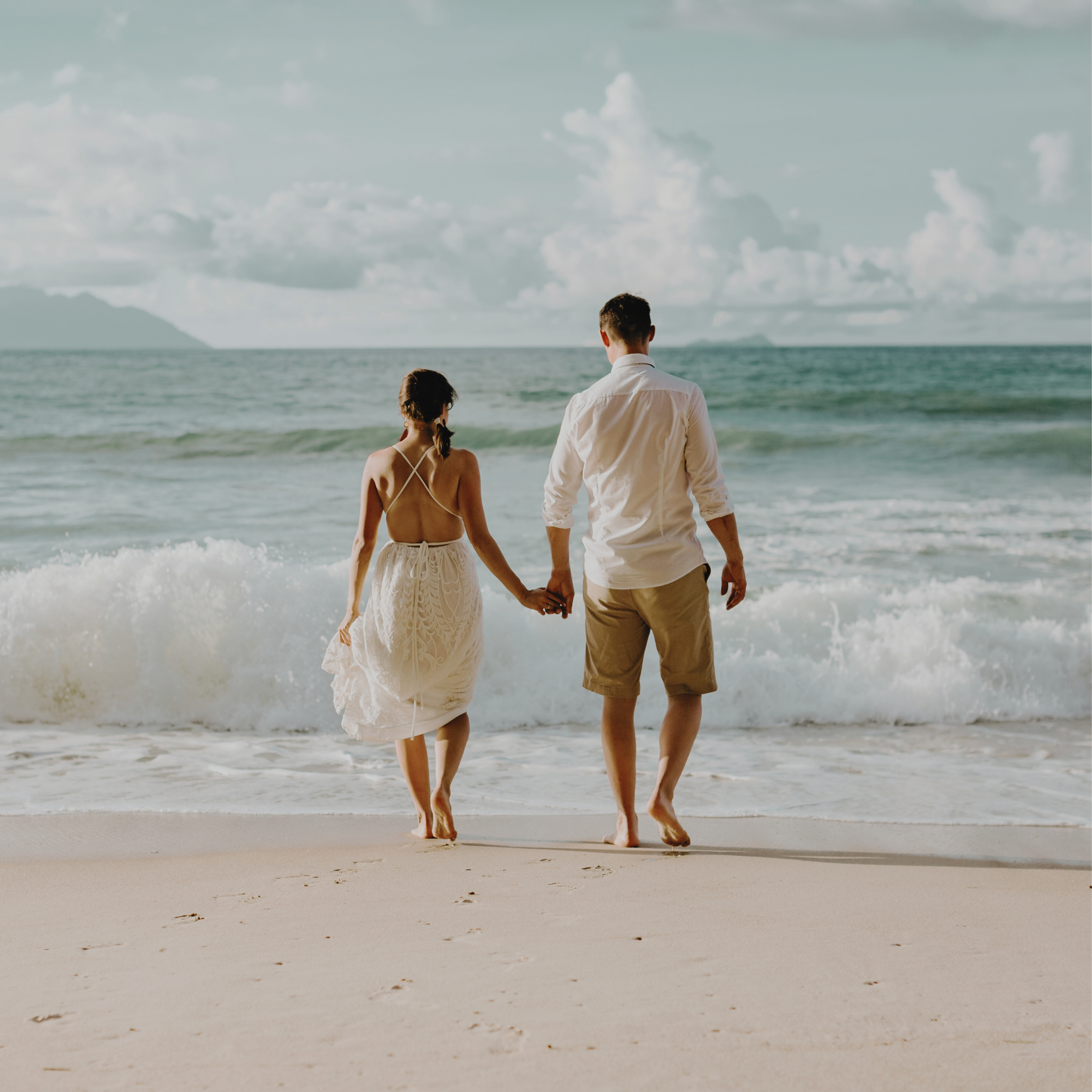 A couple walks hand in hand on the beach, with the ocean and a cloudy sky in the background.