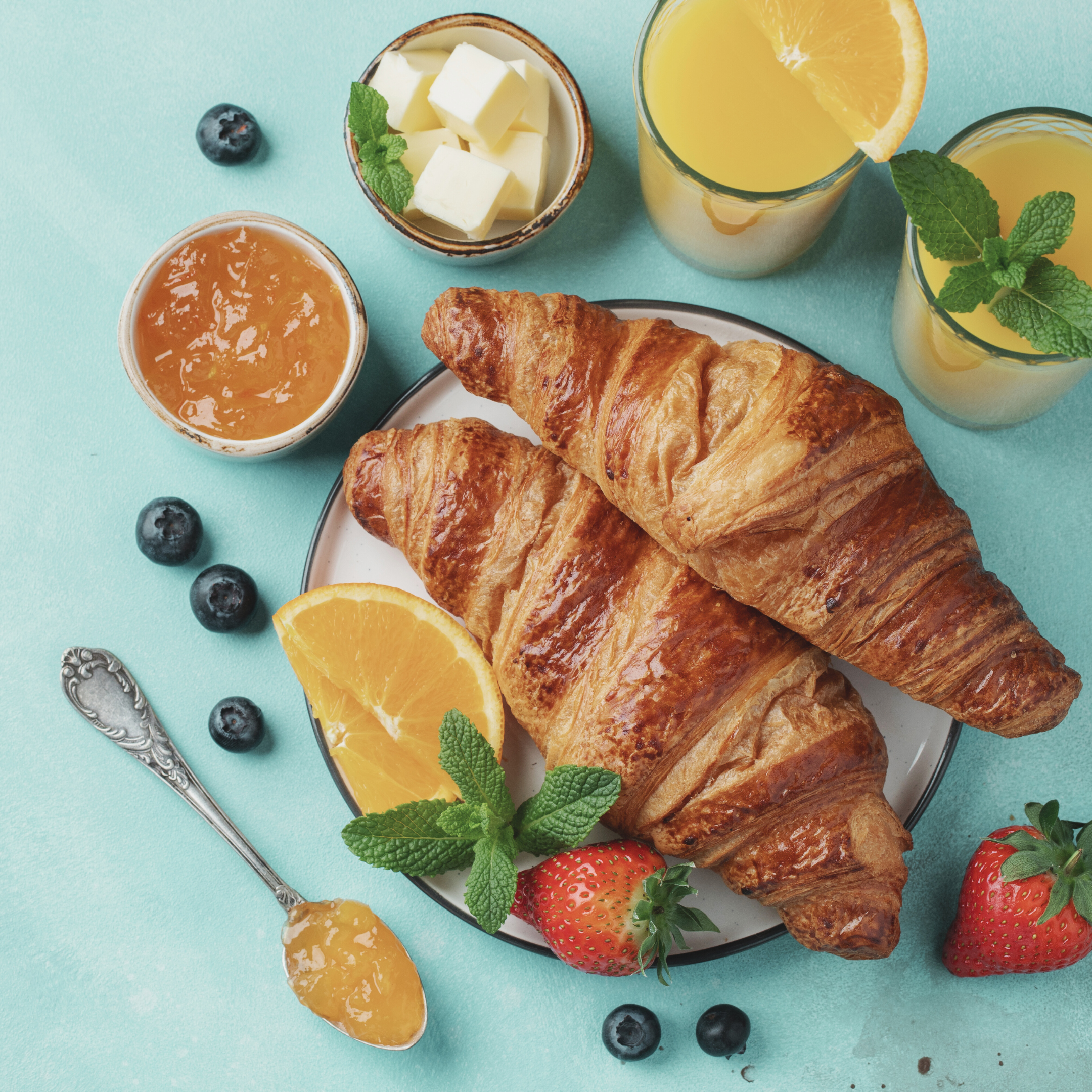 Fresh croissants, strawberries, blueberries, orange slices, and a jar of honey on a turquoise surface with glasses of orange juice and fresh mint leaves.