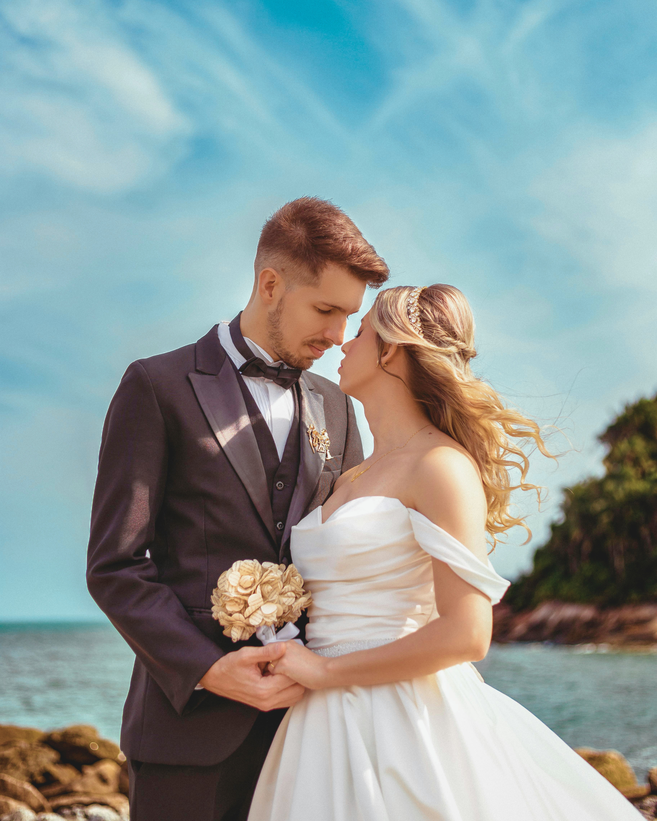 A bride and groom standing close together outdoors, forehead touching, between the ocean and a rocky shoreline, with a clear blue sky above.