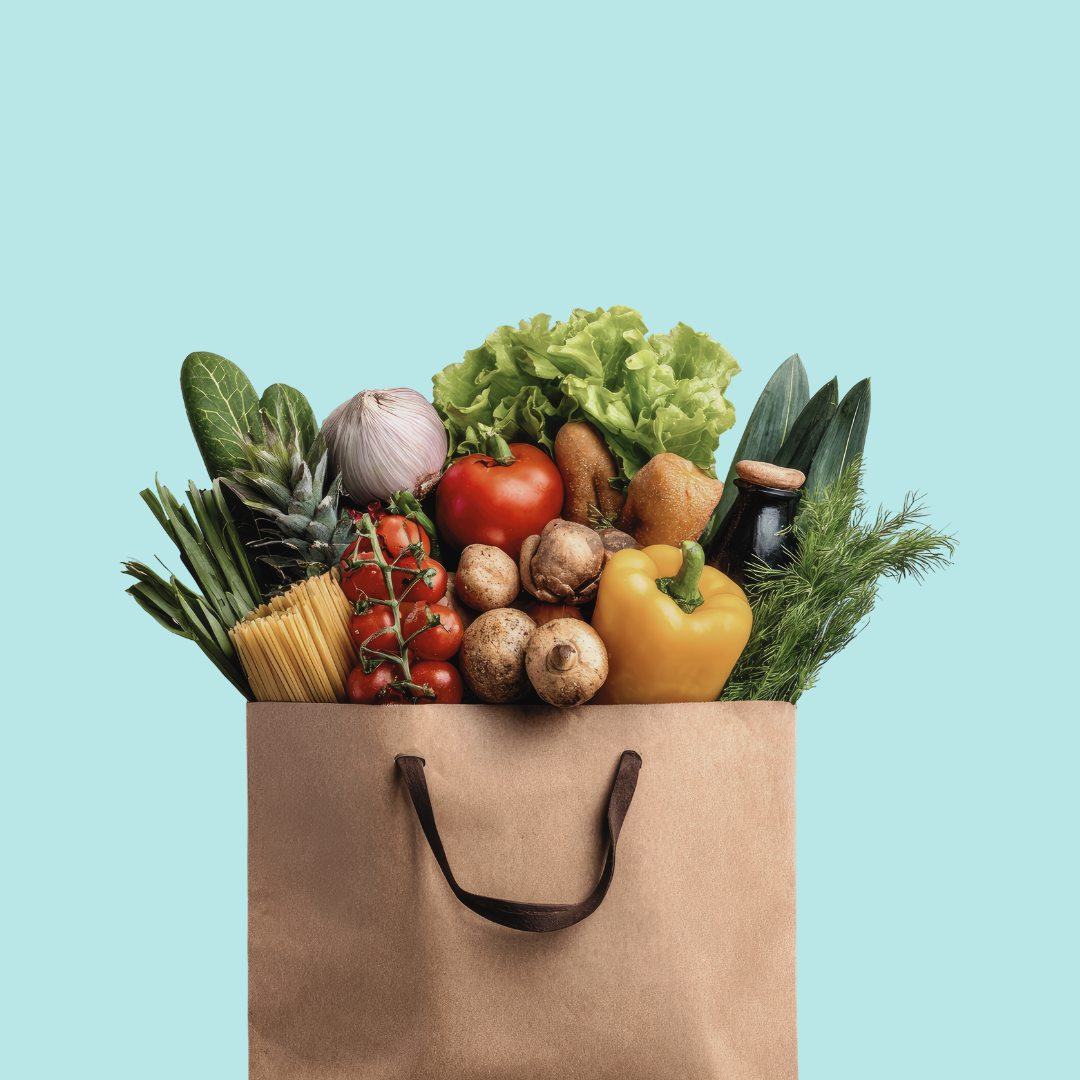 Grocery paper bag filled with vegetables including yellow bell pepper, tomatoes, lettuce, garlic, mushrooms, green onions, and other fresh produce against a blue background.