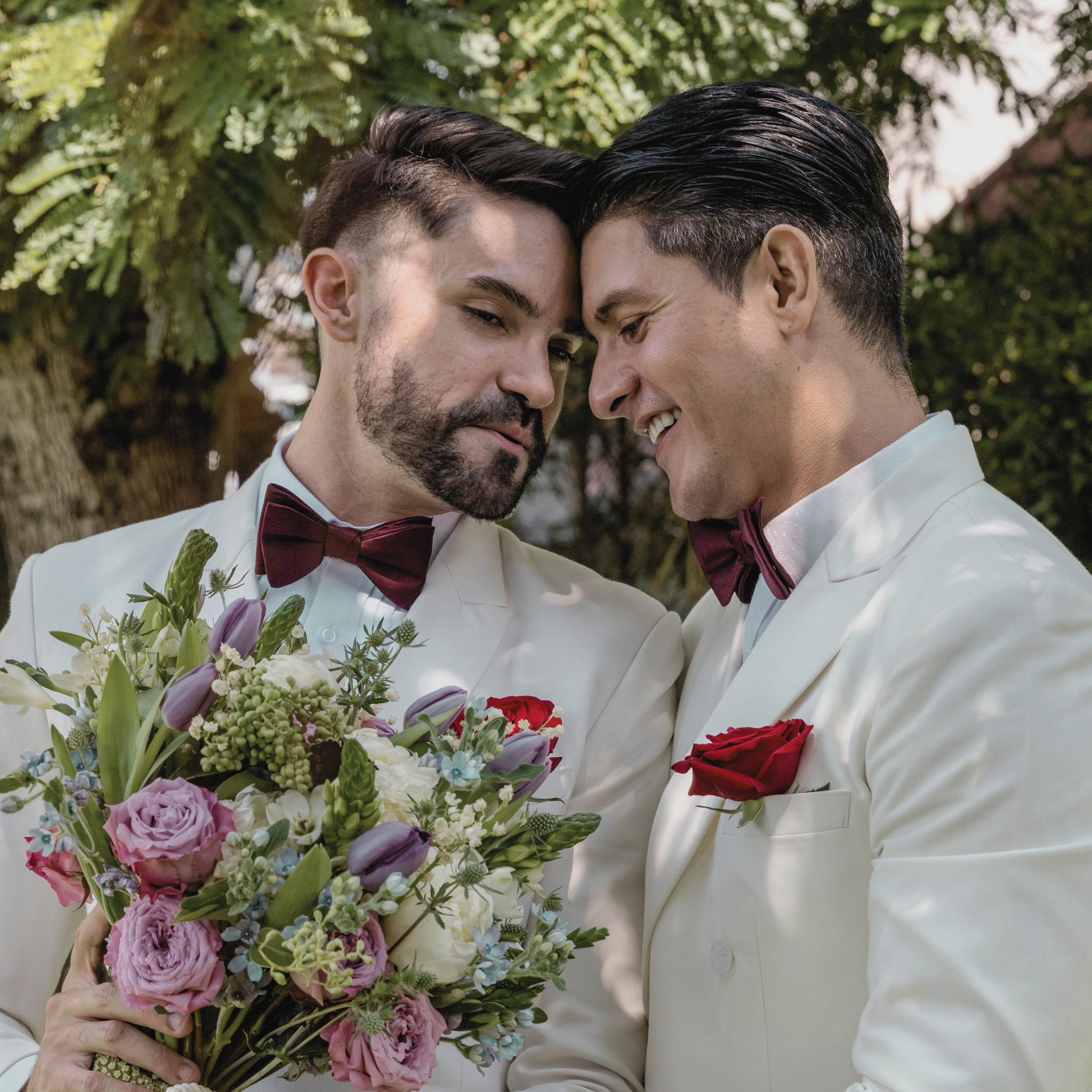 Two men dressed in white tuxedos with burgundy bow ties, standing close together with foreheads touching, smiling, and holding a large bouquet of colorful flowers including lilies, roses, and other blossoms, outdoors with green foliage in the backgro