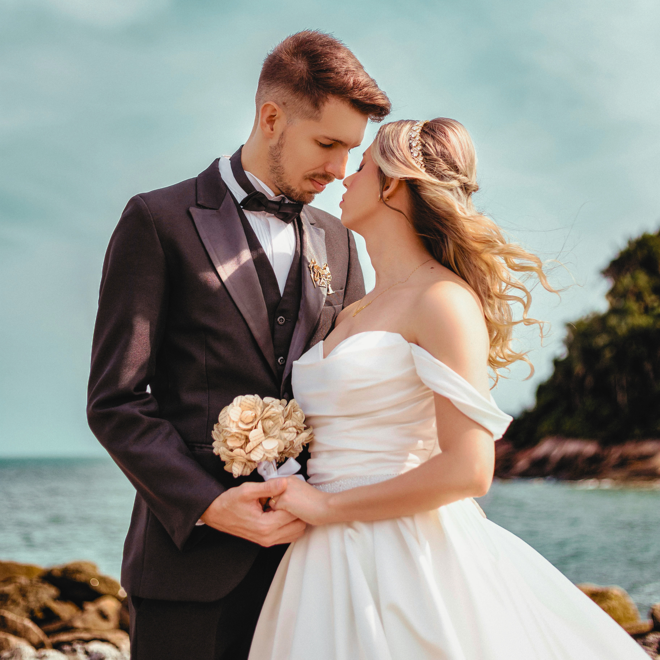 A bride and groom standing close together by the water, with their foreheads touching in an intimate moment. The groom is wearing a dark tuxedo with a bow tie, and the bride is wearing a white wedding dress and a decorative headband. The background shows a cloudy sky and a rocky landscape near the shoreline.