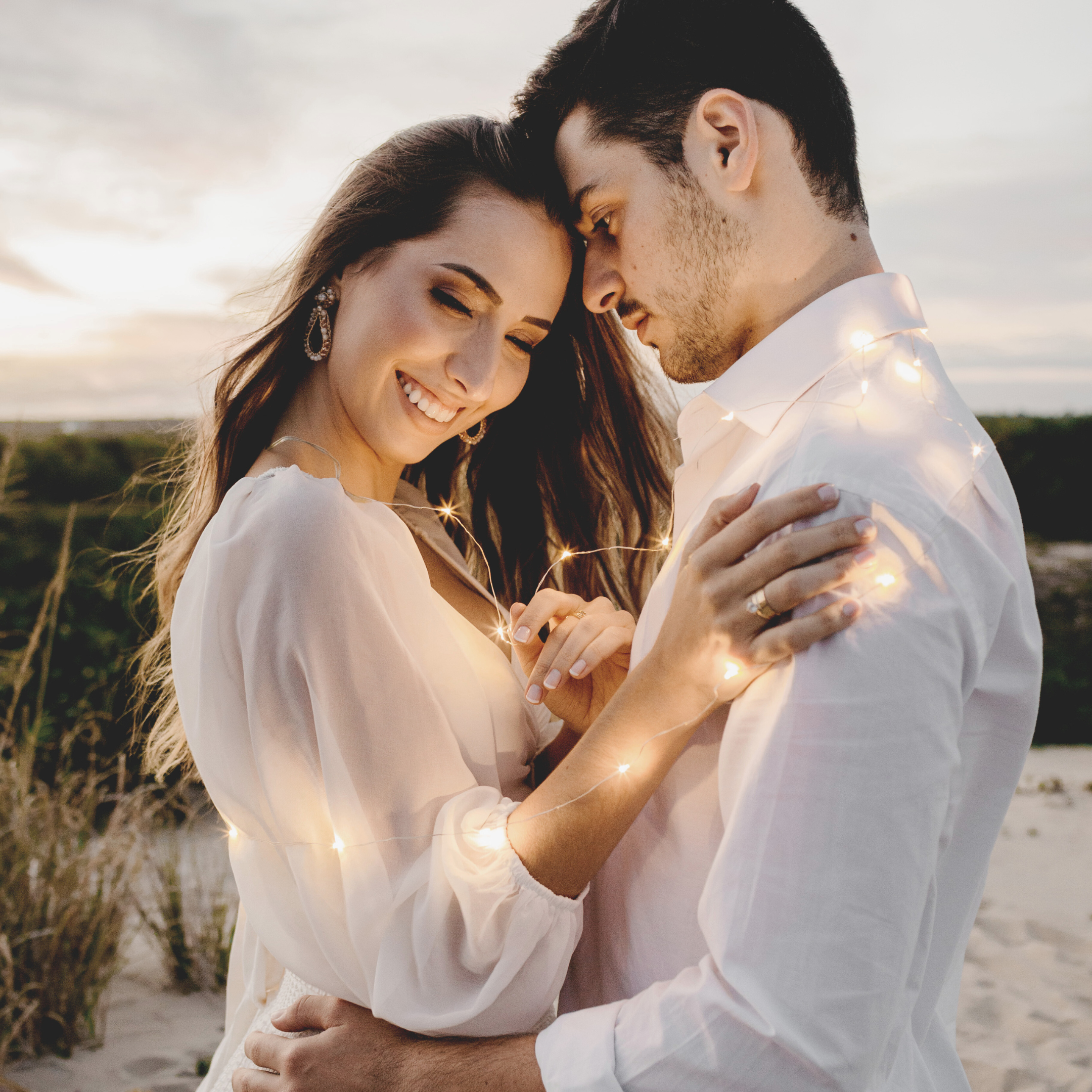 A romantic moment between a young couple, with their foreheads touching and eyes closed, outdoors during sunset, surrounded by fairy lights.