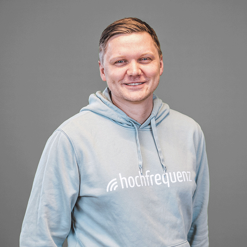 Ole trägt einen hellen Hoodie mit dem Logo von Hochfrequenz. Er steht vor einem grauen Hintergrund und lächelt in die Kamera.