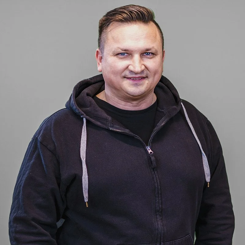 Auf dem Bild sehen wir Alex, er trägt eine dunkle Hoodie Jacke, steht vor einem grauen Hintergrund und lächelt in die Kamera.