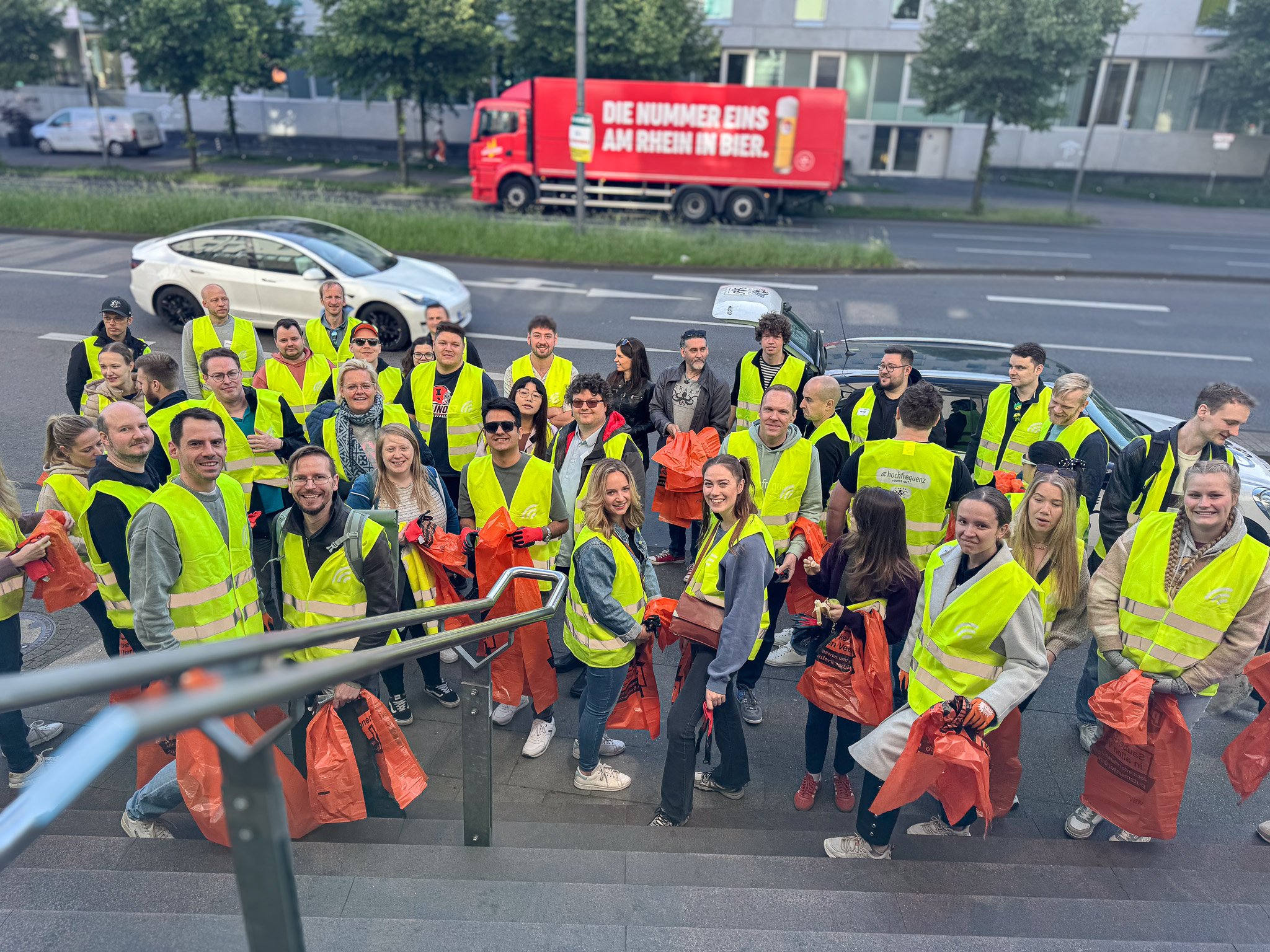 Gruppe von Menschen in gelben Sicherheitswesten stehen auf Treppe im Freien, halten orange Taschen, im Hintergrund Straße mit Autos und einem roten Truck mit Werbung.