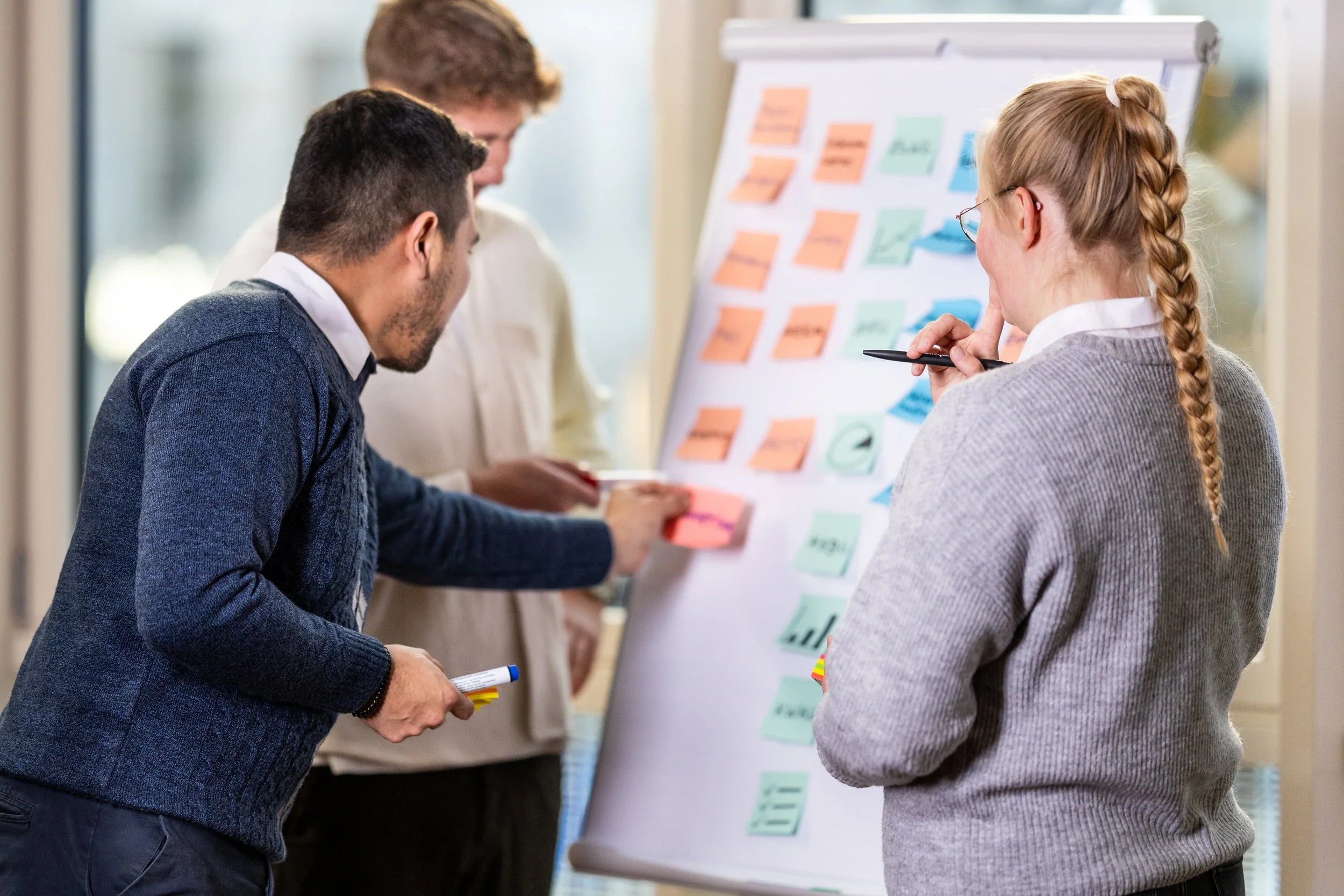 Drei Personen arbeiten gemeinsam an einem Whiteboard mit bunten Haftnotizen, die bei einer Gruppen Brainstorming oder Planungssitzung verwendet werden.