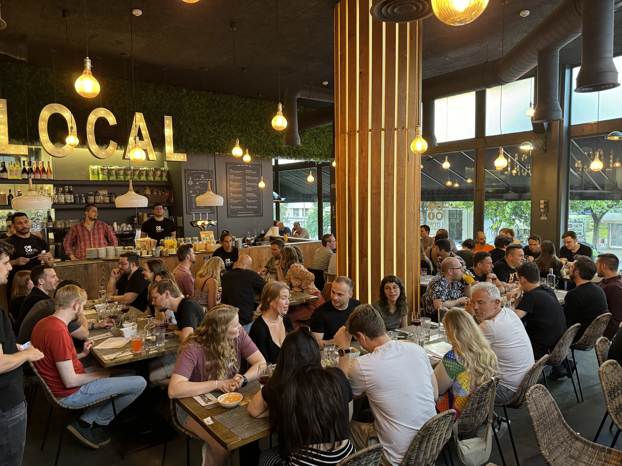 eine große Gruppe an Leuten ist an verschiedenen Tischen verteilt im Restaurant. Alle unterhalten sich und lachen zusammen. An einigen Tischen steht schon Essen auf dem Tisch. 