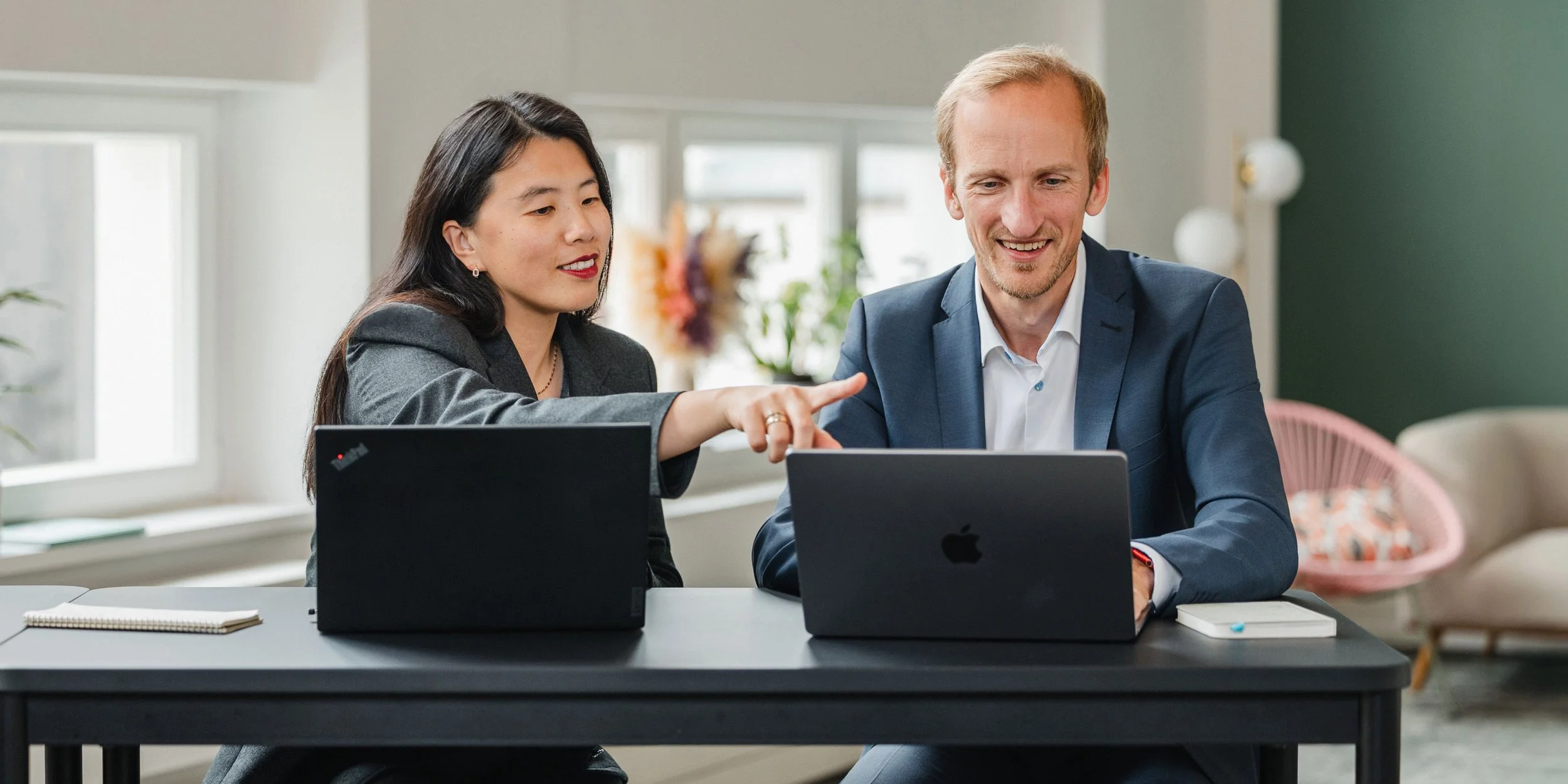 Ein Mann und eine Frau sitzen zusammen an einem Tisch, beide nutzen Laptops und unterhalten sich.