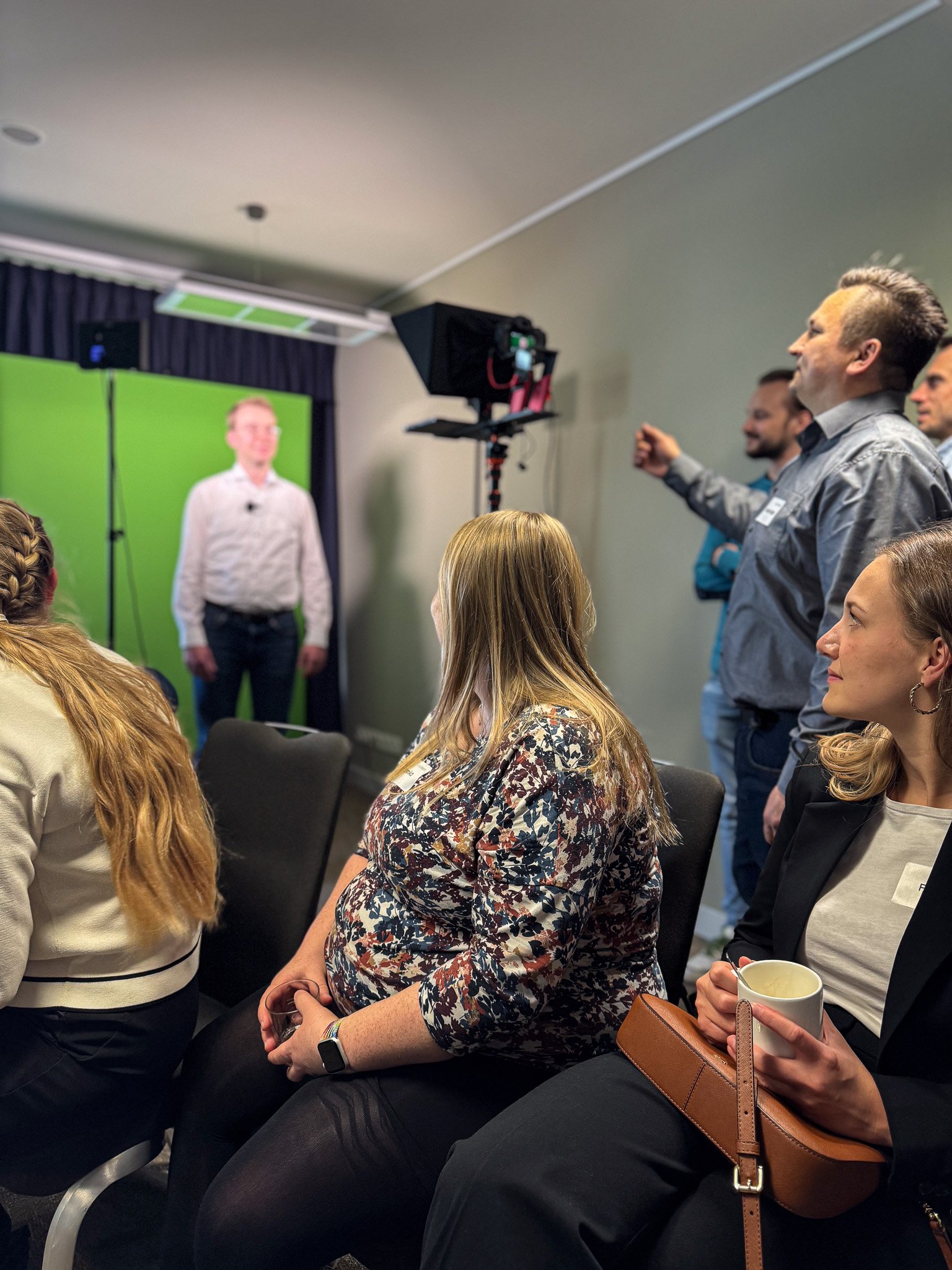 Eine Filmaufnahme von HF erklärt wird hier simuliert. Man sieht einen Mann vor der Kamera und mehrere hinter der Kamera. Vorne im Bild sitzen drei Frauen, die das ganze beobachten. 