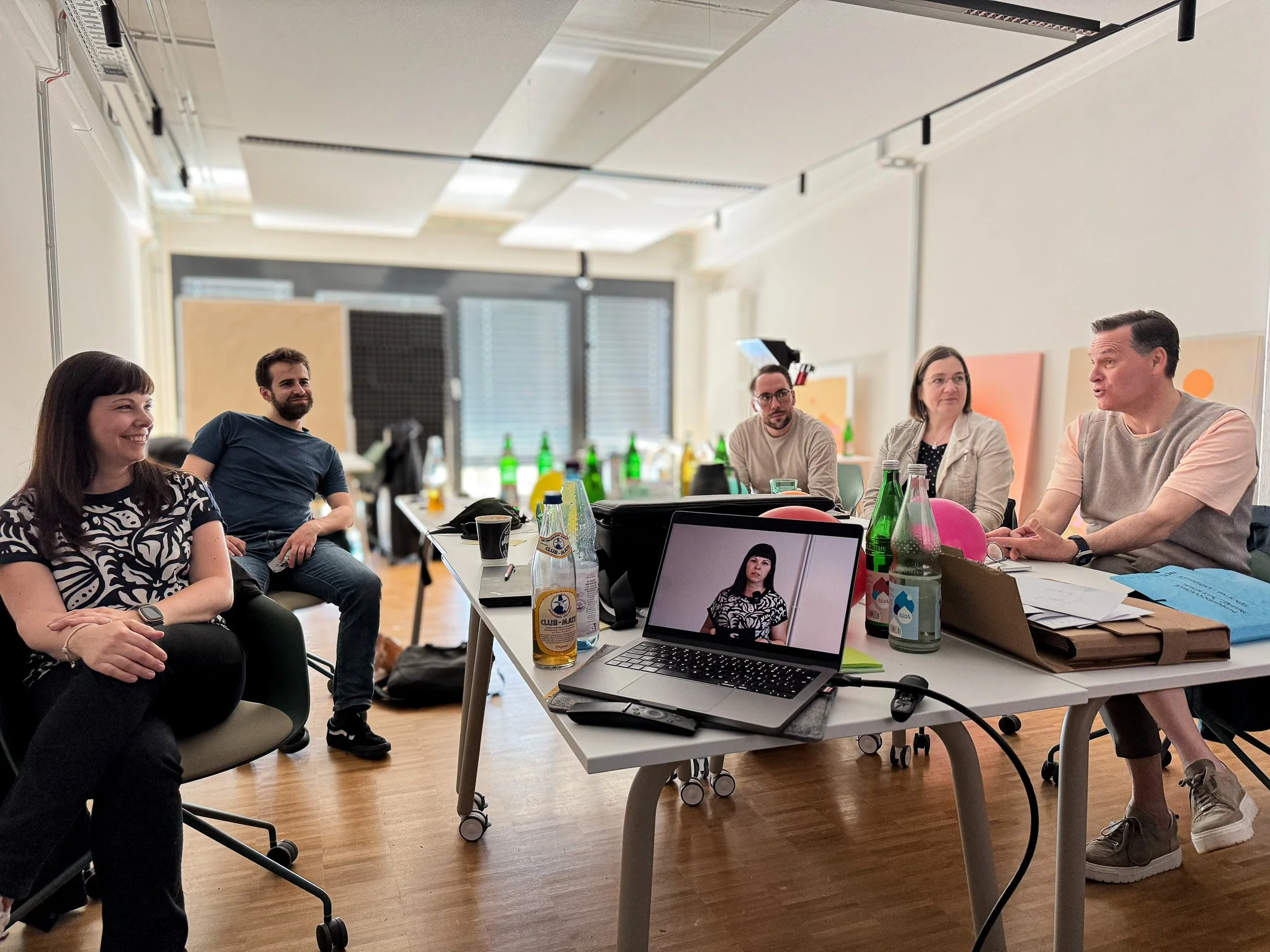 Fünf erwachsene Personen sitzen in einem Besprechungsraum um einen Tisch. Es gibt verschiedene Getränke und Dokumente auf dem Tisch. Auf einem Laptop ist ein Bild einer Frau zu sehen, die auch im Raum sitzt. Die Atmosphäre wirkt entspannt, einige Per