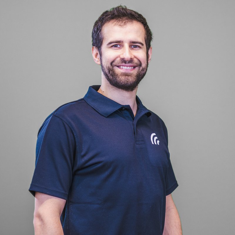 Lukas steht vor einem grauen Hintergrund. Er trägt ein dunkelblaues Poloshirt mit dem Hochfrequenz Logo.