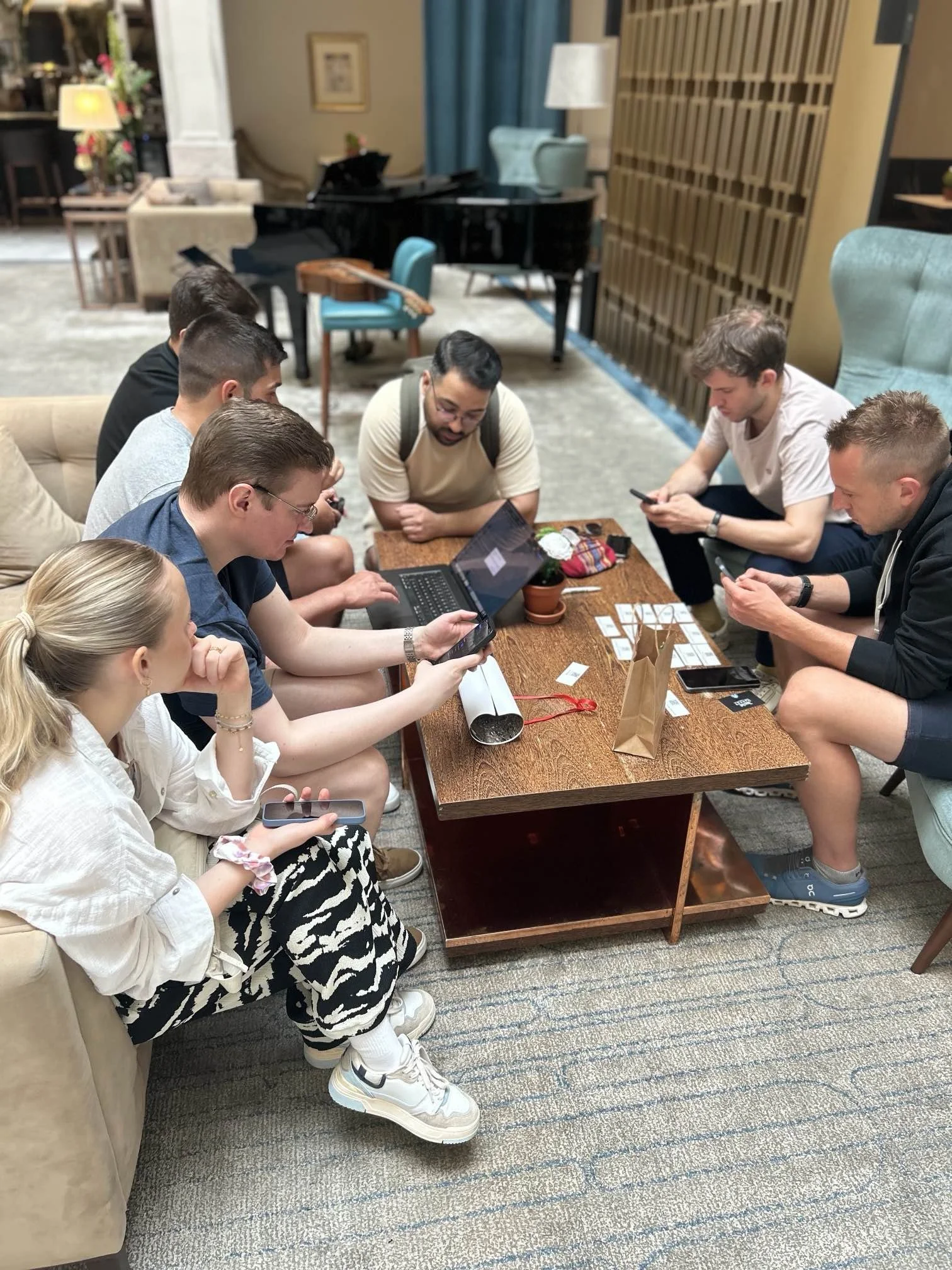 Sieben Leute sitzen zusammen an einem kleine Tisch und betrachten  ein Rätsel. Ein Teil der Gruppe versucht es am PC zu lösen und ein anderer Teil der Gruppe hat das Handy in der Hand. 