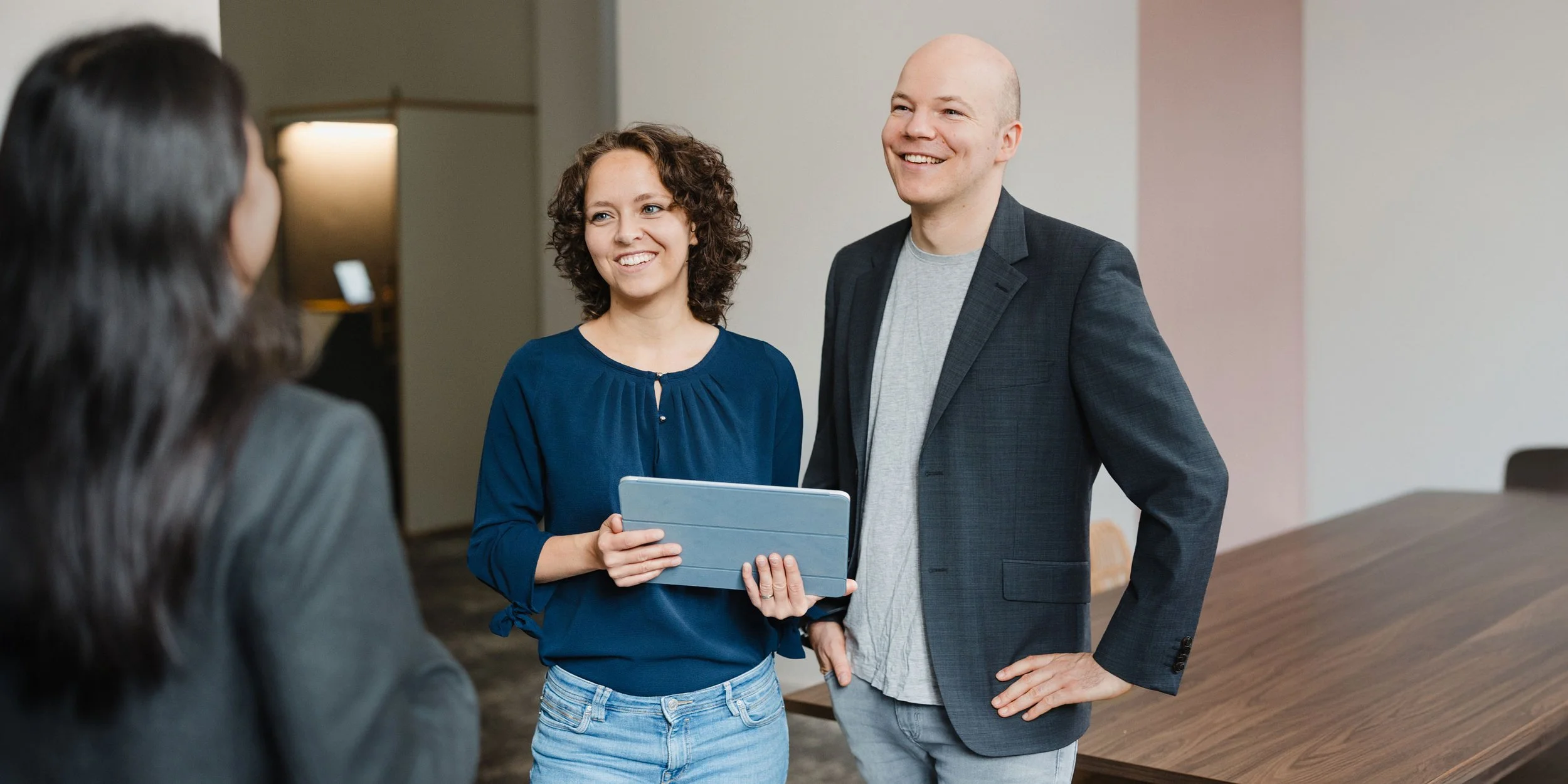 Drei Personen unterhalten sich in einem modernen Büro, eine hält ein Tablets, alle lachen und wirken entspannt.