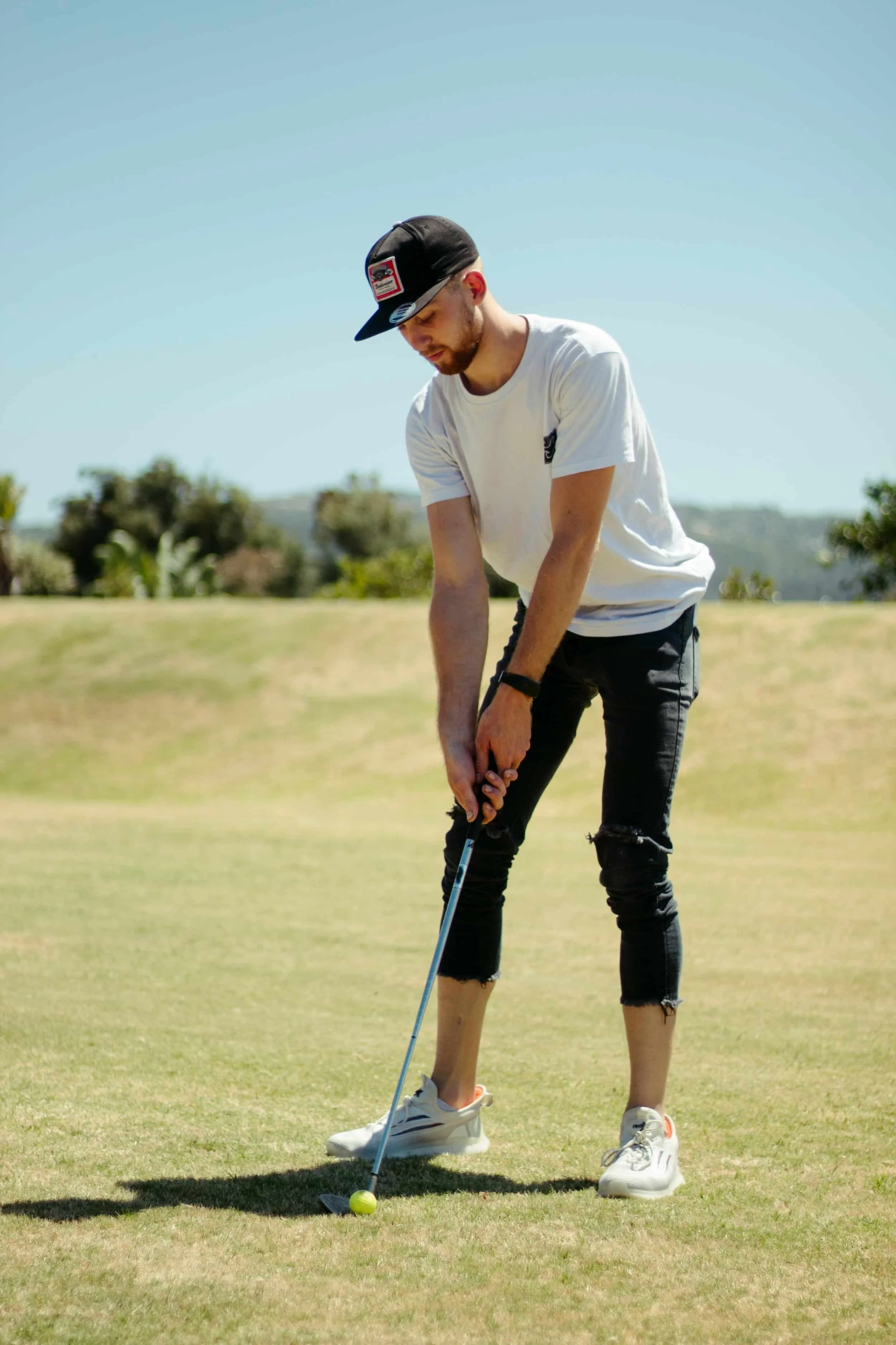 A man wearing a white t-shirt, ripped black jeans, white sneakers, and a black cap is preparing to hit a golf ball on a golf course.