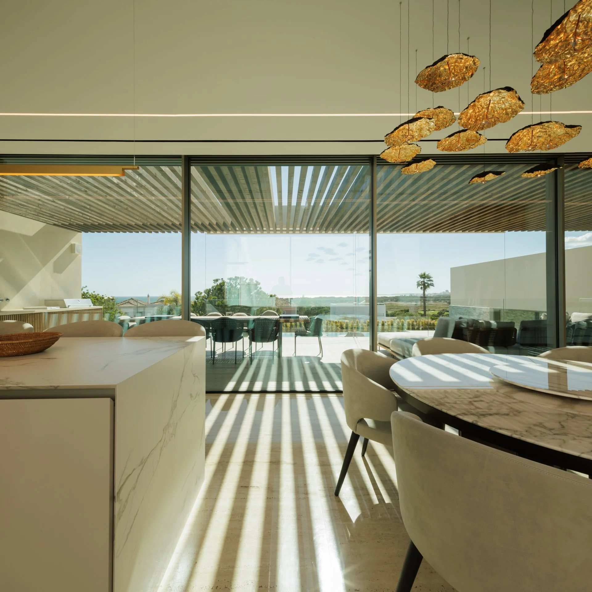 Luxury white and marble kitchen with a matching dining table and grey dining chairs under a statement lighting piece, looking out onto a balcony with breathtaking views