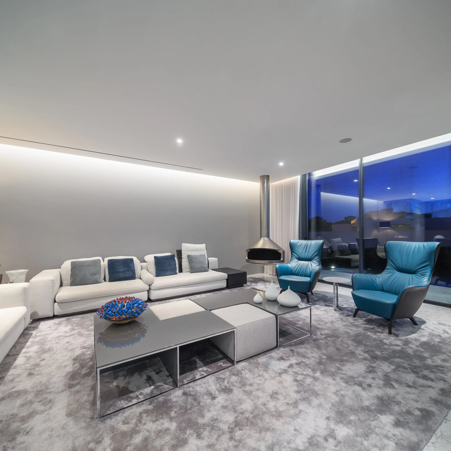 White and blue themed living room project by A+ Interiors with modern furniture and a suspended fireplace, matching the stunning backdrop of the early evening sky 