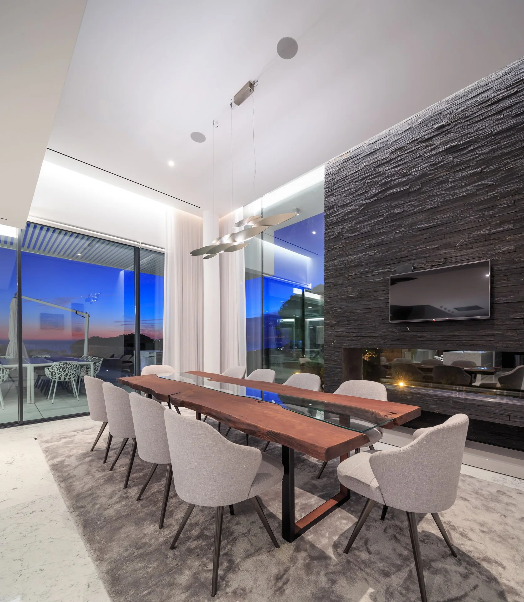Stunning dining space at sunset, with a dark stone wall and integrated fireplace, wood and glass statement dining table, and simple grey dining chairs 
