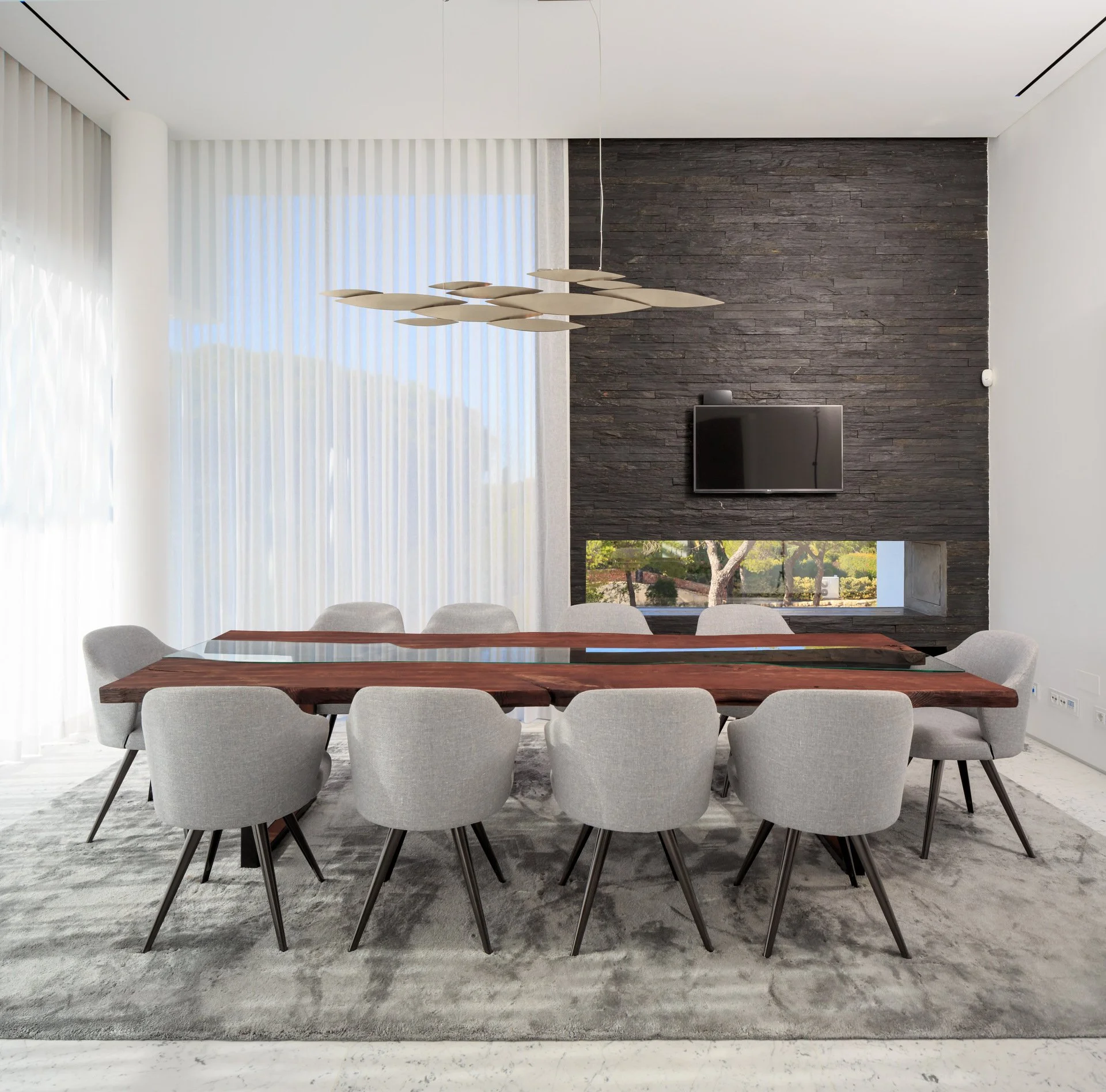 Stunning dining space with a dark stone wall and integrated fireplace, wood and glass statement dining table, simple grey dining chairs and a contemporary lighting piece