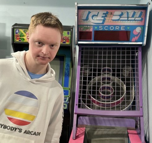 A young man with red hair and a white ANEWAY hoodie stands next to a ski ball arcade game