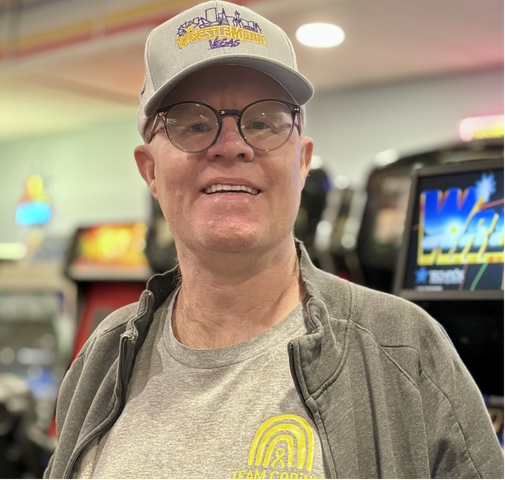 A man is wearing a gray t-shirt with a gray hat, a Wrestlemania baseball cap and glasses. He is standing in front of aracade games and smiling to the camera