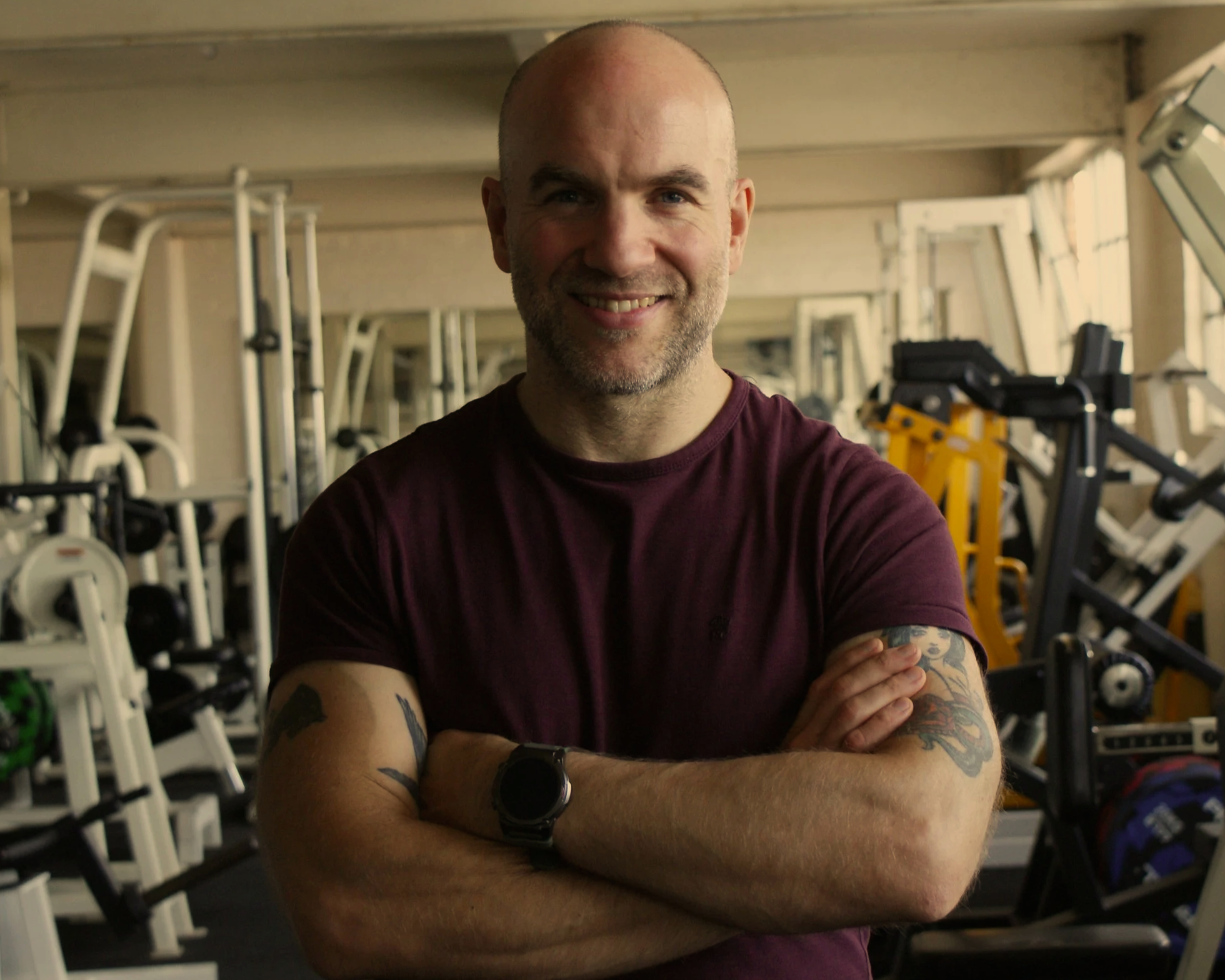 A man with tattoos on his arms, wearing a maroon shirt and a watch, standing with arms crossed in a gym with exercise equipment.