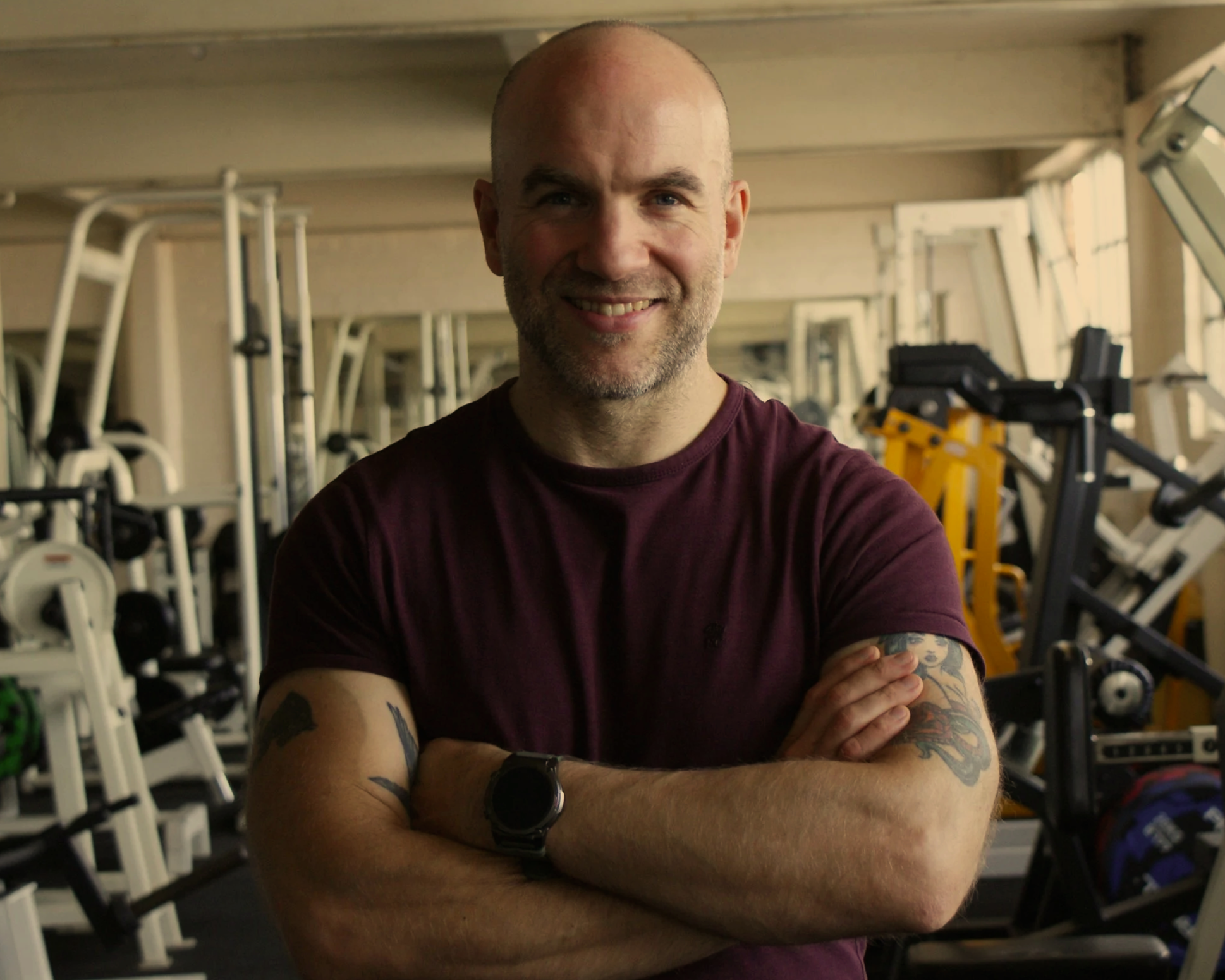 A friendly male personal trainer with a warm smile, wearing a maroon t-shirt and a black smartwatch, standing poised and proud with crossed arms in a gym filled with exercise equipment, ready to motivate