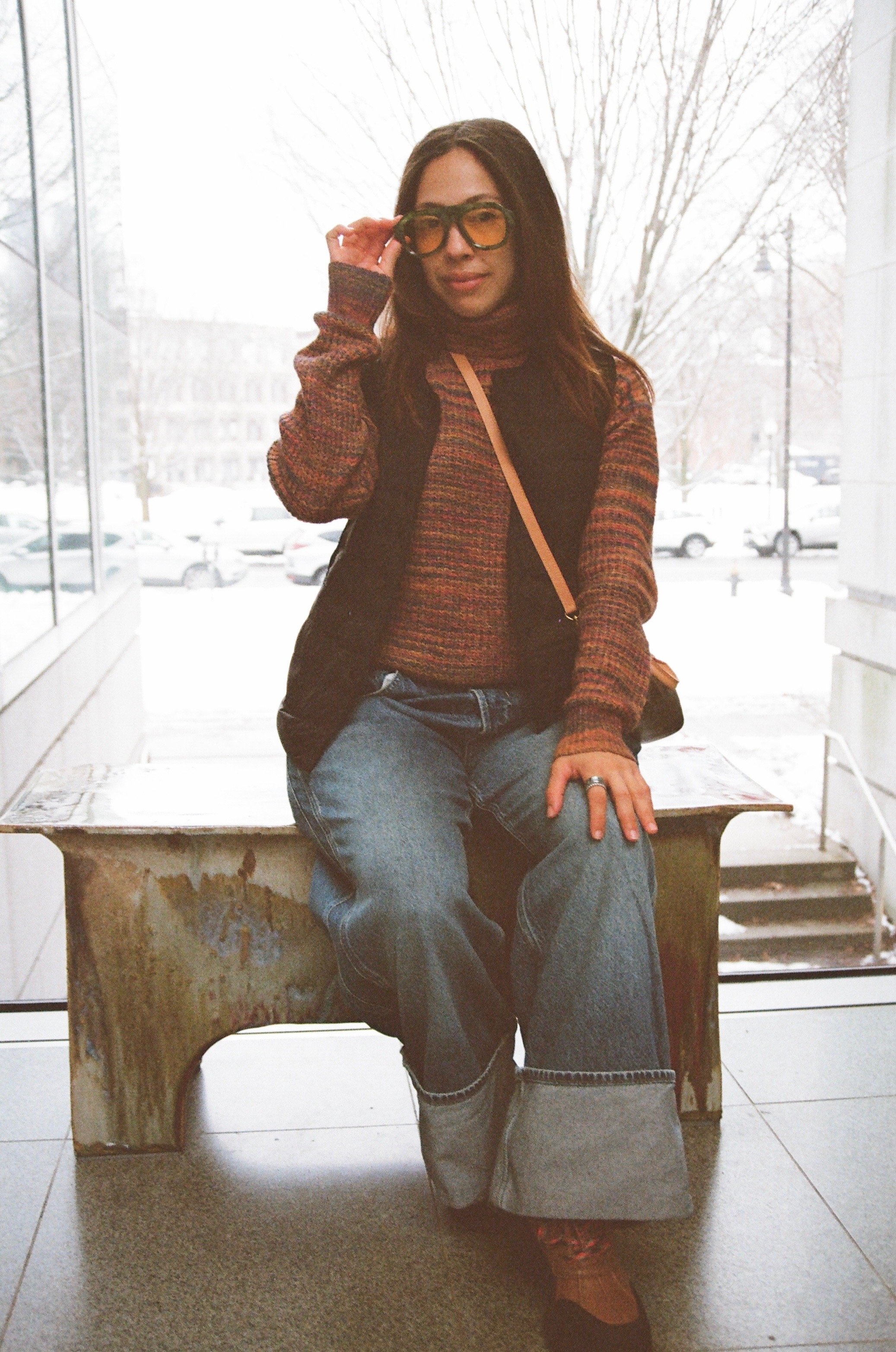 Woman seated on a wooden bench indoors, adjusting sunglasses, wearing a multicolored sweater, black vest, wide-leg jeans, and glasses, with a snowy outdoor scene visible through large windows behind her.