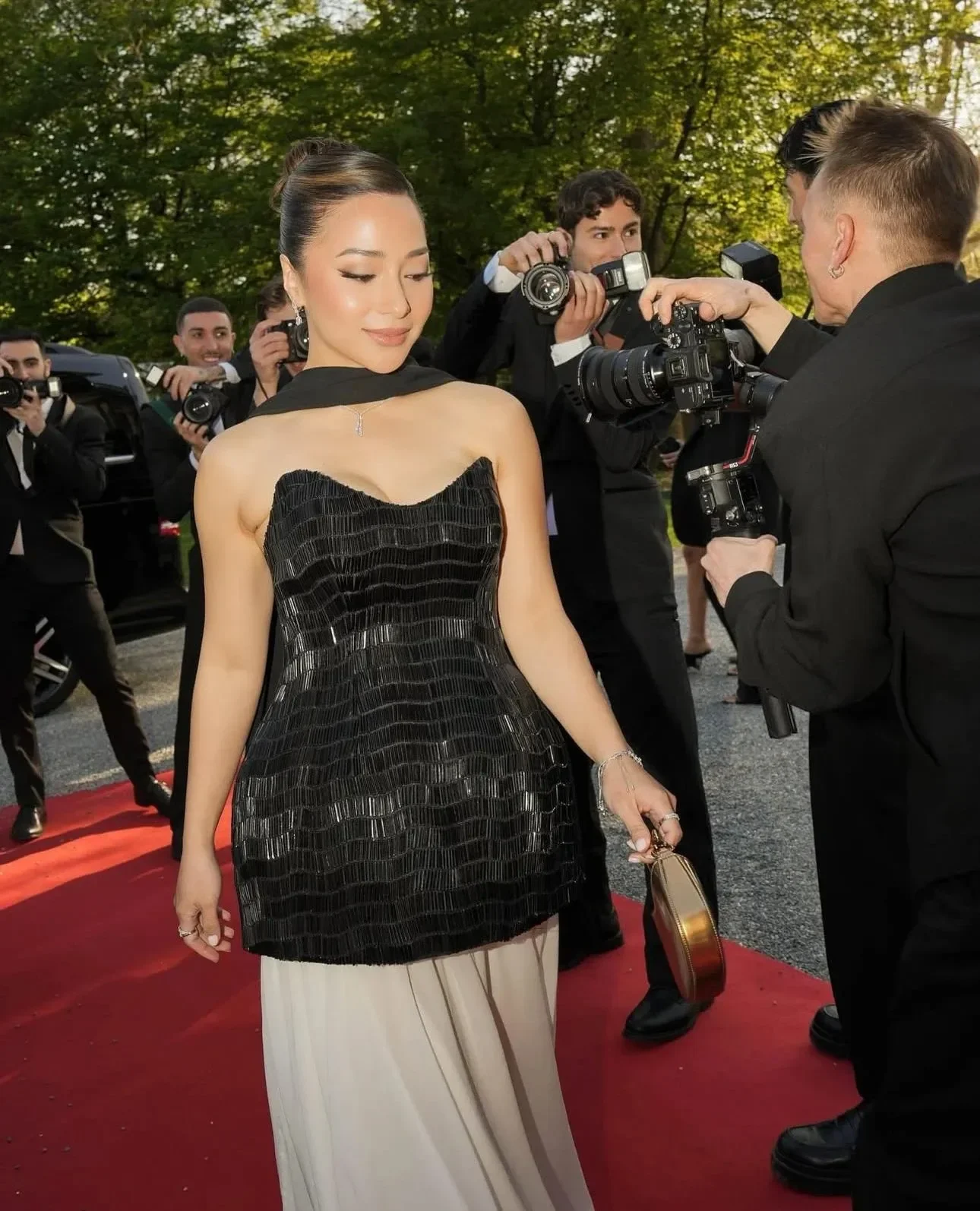 A woman in an elegant black strapless dress and beige skirt walks on a red carpet surrounded by photographers.