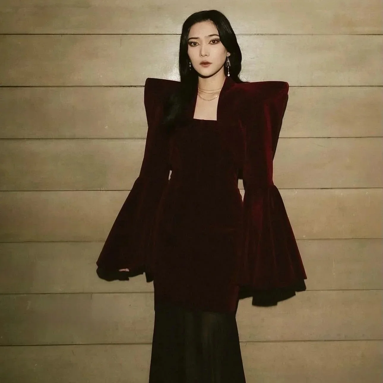 A woman with dark hair wearing a dark red velvet dress with exaggerated shoulder pads, standing against a wooden paneled wall.
