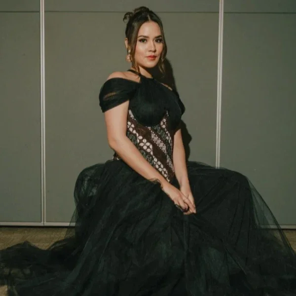 A woman sitting on the floor wearing an elegant black gown with lace and satin details, and off-shoulder puff sleeves, in front of a plain wall.