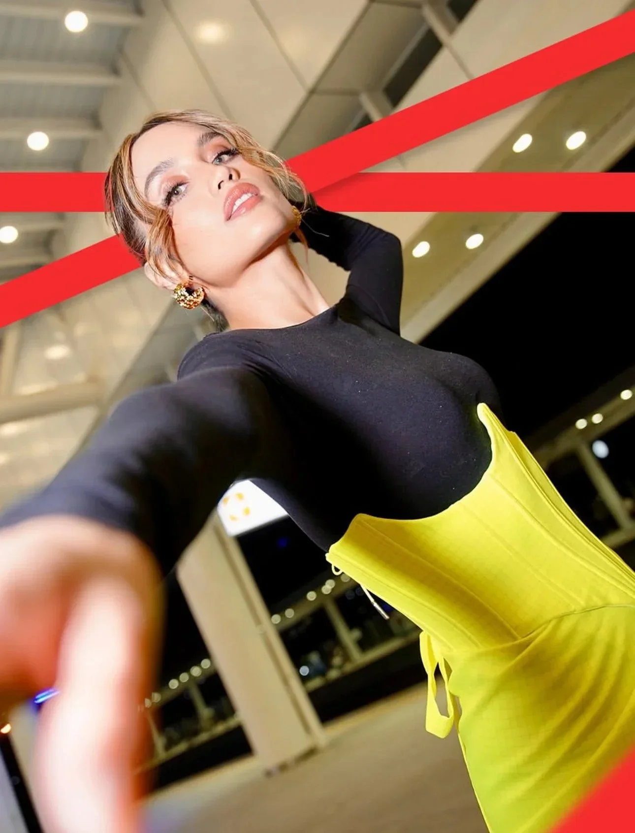 A woman taking a selfie at an airport with red lines crossing through the image, wearing a black top and a yellow skirt.