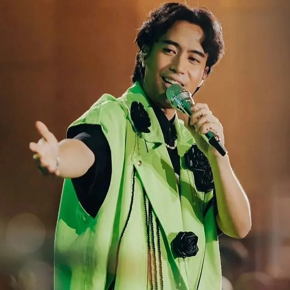 A singer performing on stage, wearing a green jacket with black accessories, holding a microphone, and extending his hand towards the audience.