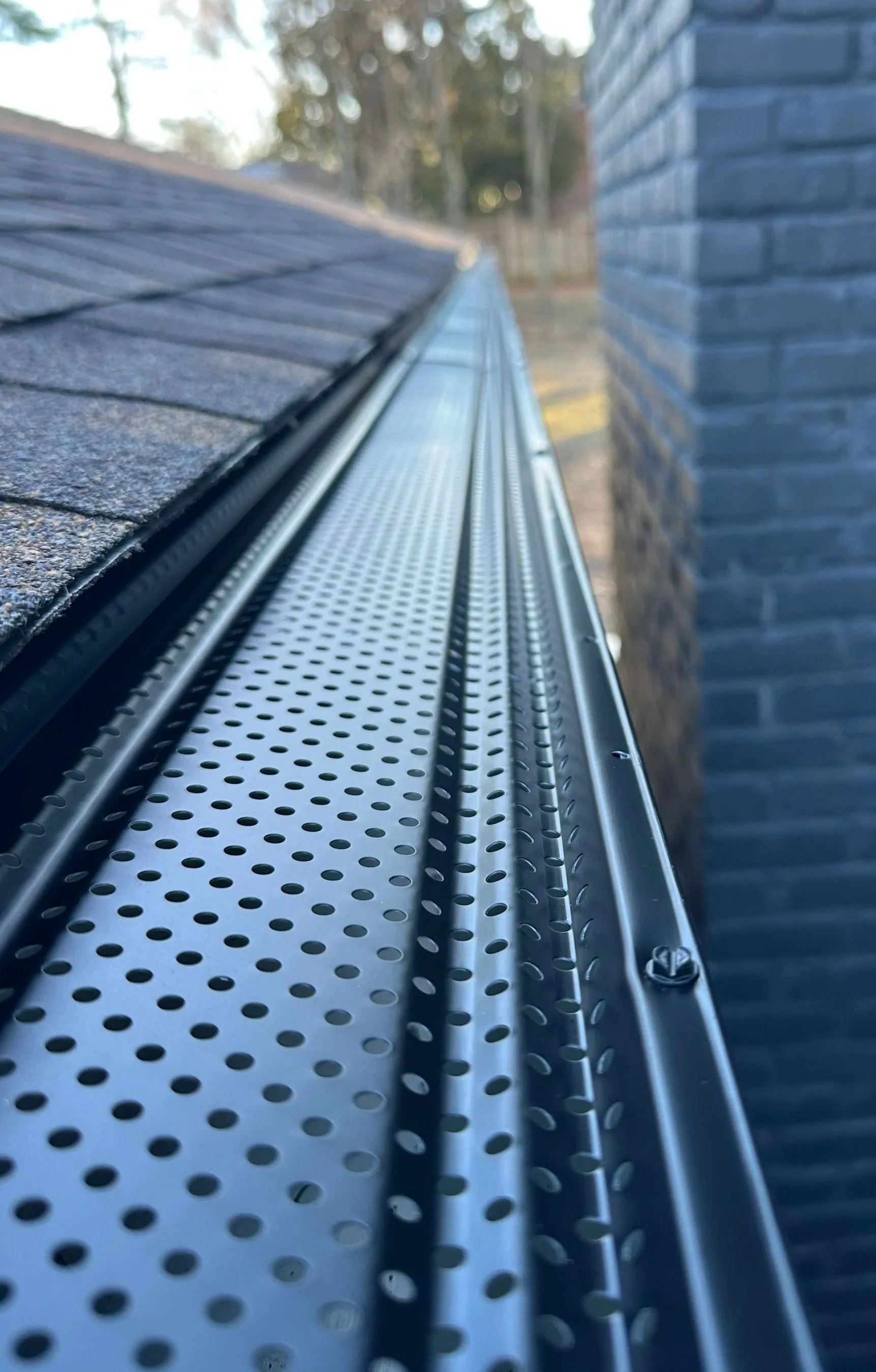 Close-up of metallic gutter guards with perforations, mounted along the edge of a shingled roof next to a brick chimney, with trees in the background.