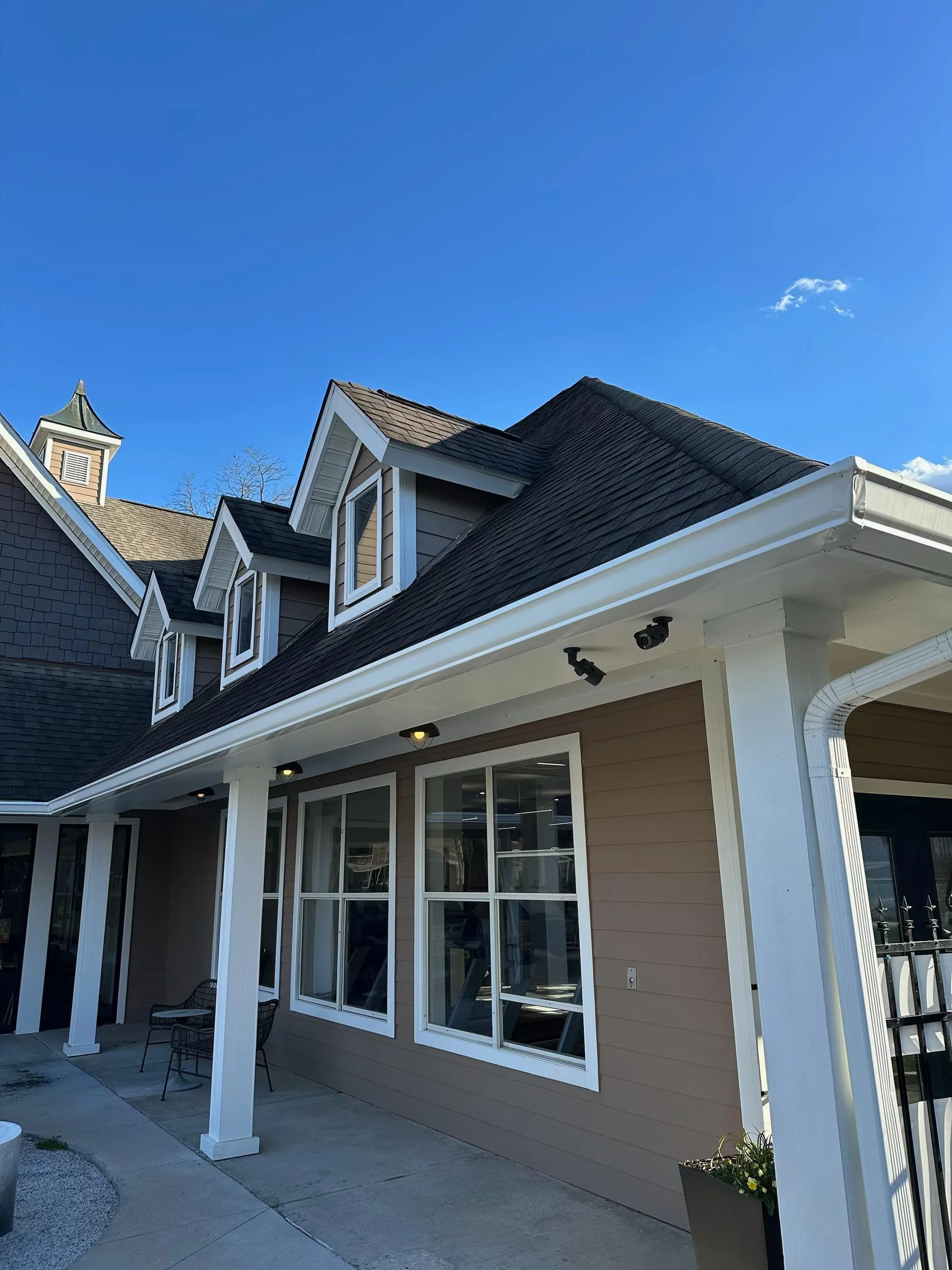 Exterior view of a beige house with white trim, three dormer windows on the roof, and a covered patio with seating and lighting, under a blue sky.