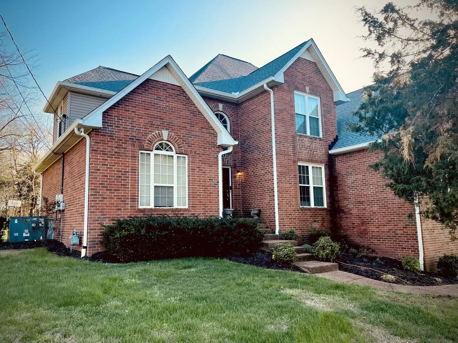 Front view of a large brick house with a well-kept lawn and bushes, new gutters, multiple windows, and a small staircase leading to the front door, on a clear day.