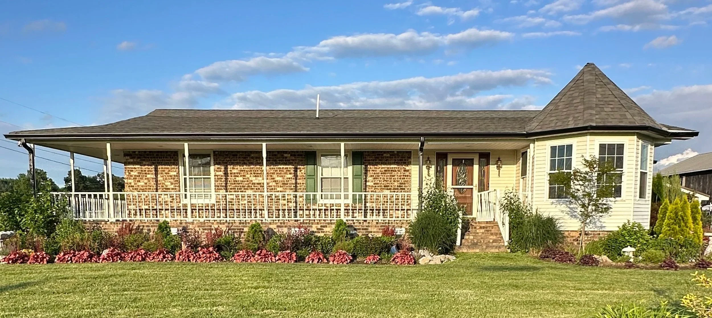 A house with a brick and yellow exterior, new gutters, a porch with railing, and a landscaped garden with colorful flowers in front.