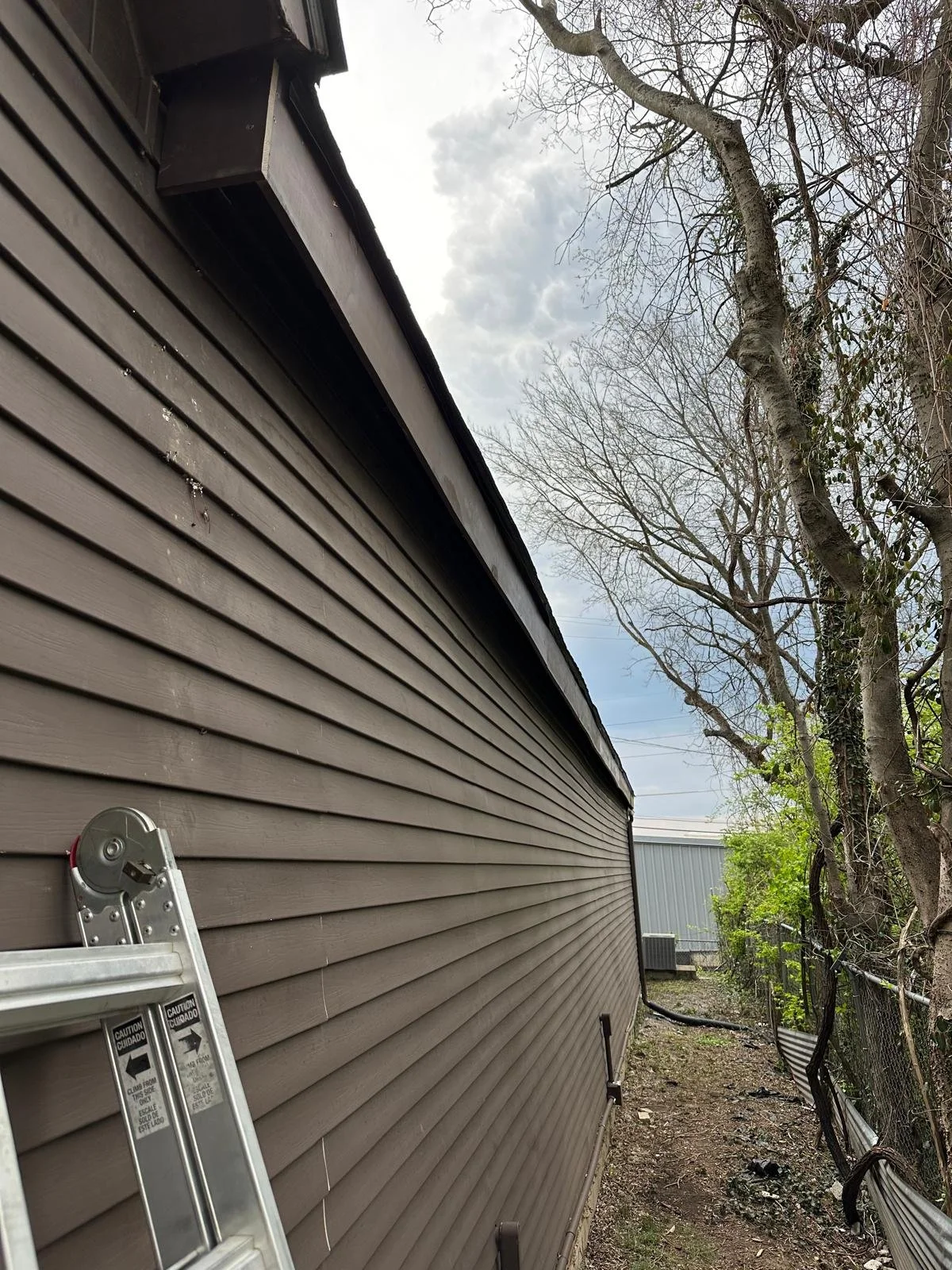 Side of a house with brown siding, a metal ladder leaning against it, trees with bare branches, and a cloudy sky.