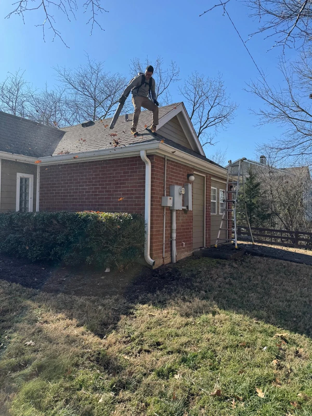 A person standing on the roof of a house, holding a leaf blower, with a ladder leaning against the house and fallen leaves on the roof.