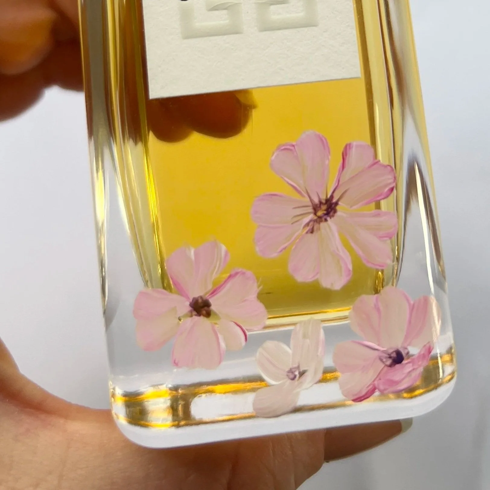 A glass vase with pink flowers submerged in yellow liquid, held by a hand against a white background.