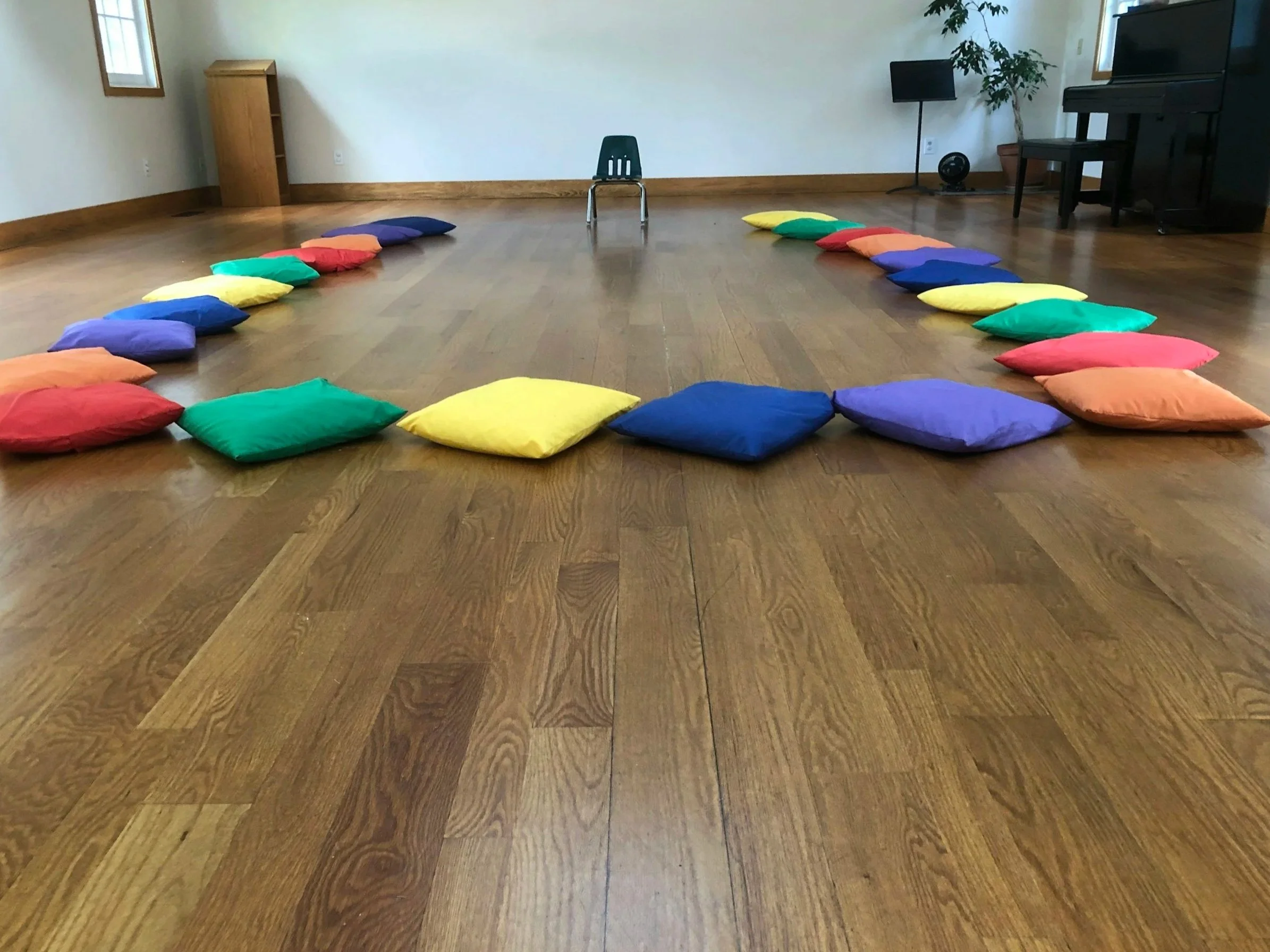 Colorful pillows arranged in a large circle on a wooden floor in a spacious room with minimal furniture.