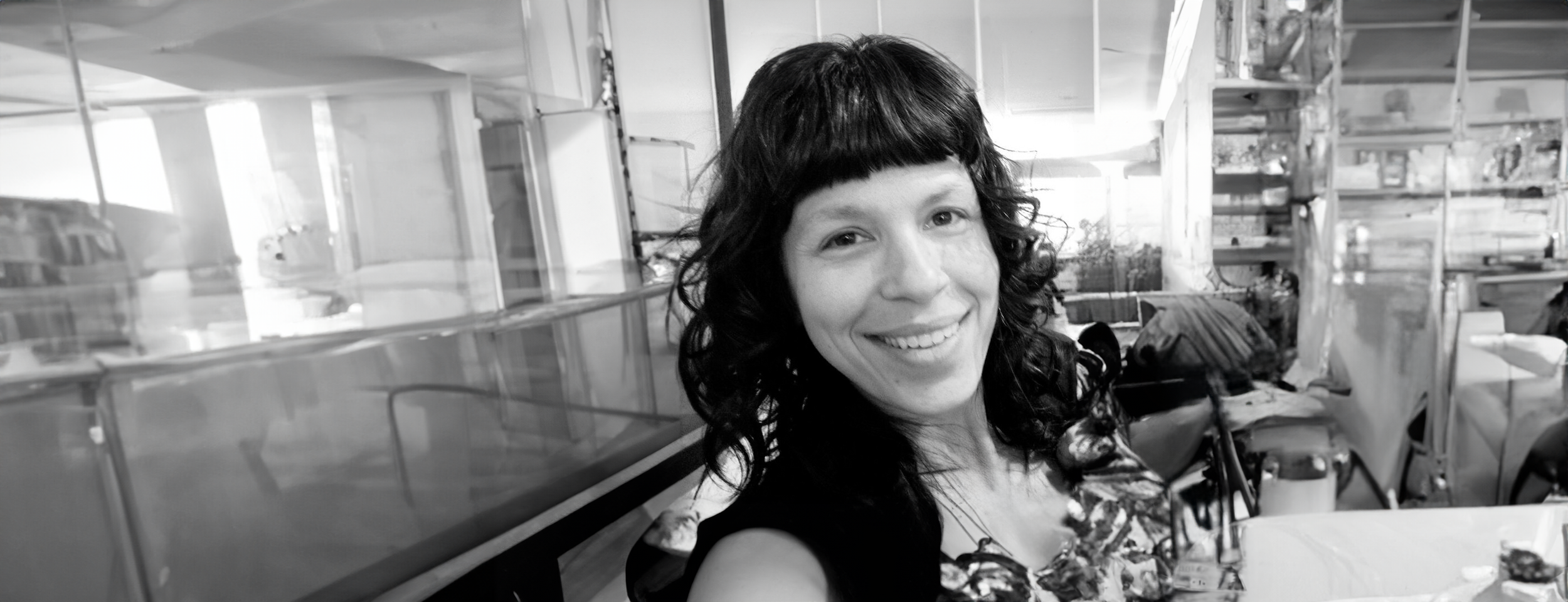 Black and white photo of a smiling person with long curly hair, standing indoors with shelves and various items in the background.