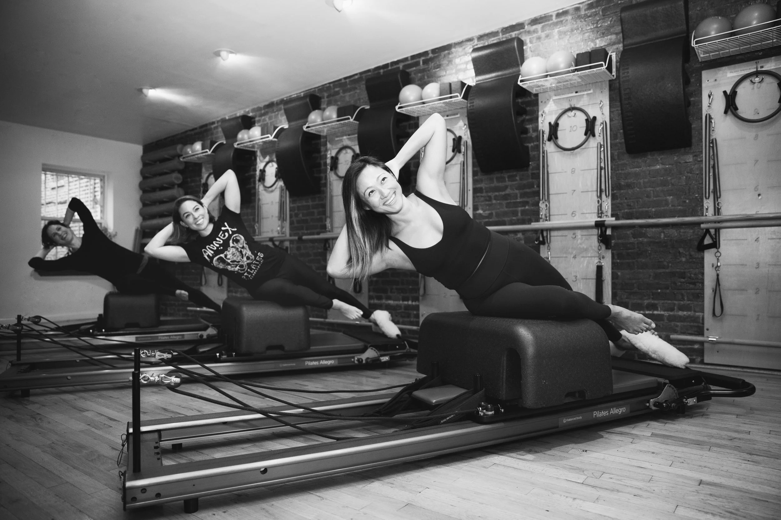 Three women exercising on Pilates reformer machines in a fitness studio, with brick walls and exercise equipment in the background.