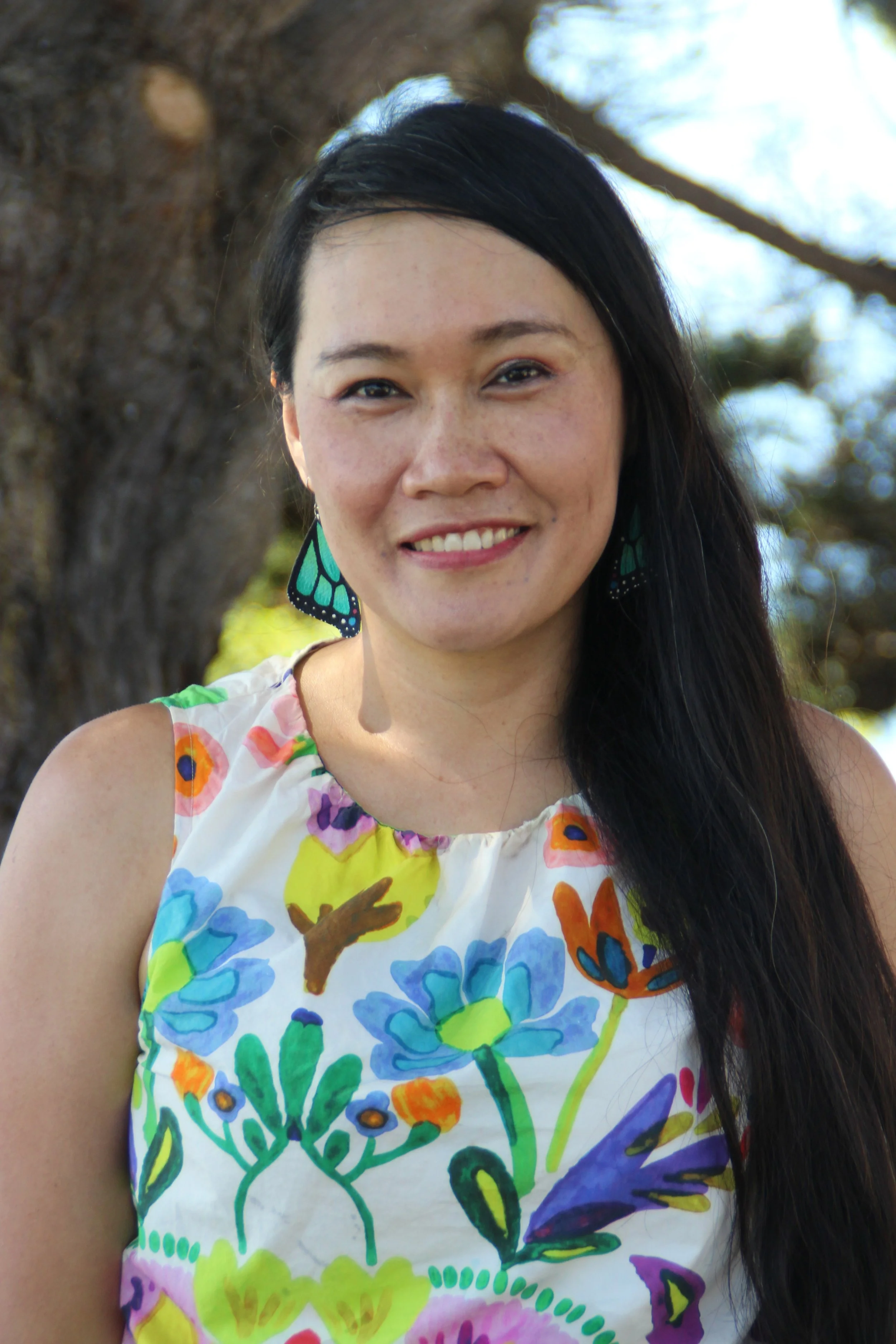 Close-up photo of a woman with long black hair, smiling, wearing butterfly-shaped earrings and a colorful sleeveless top with floral and butterfly patterns, standing outdoors near a large tree.