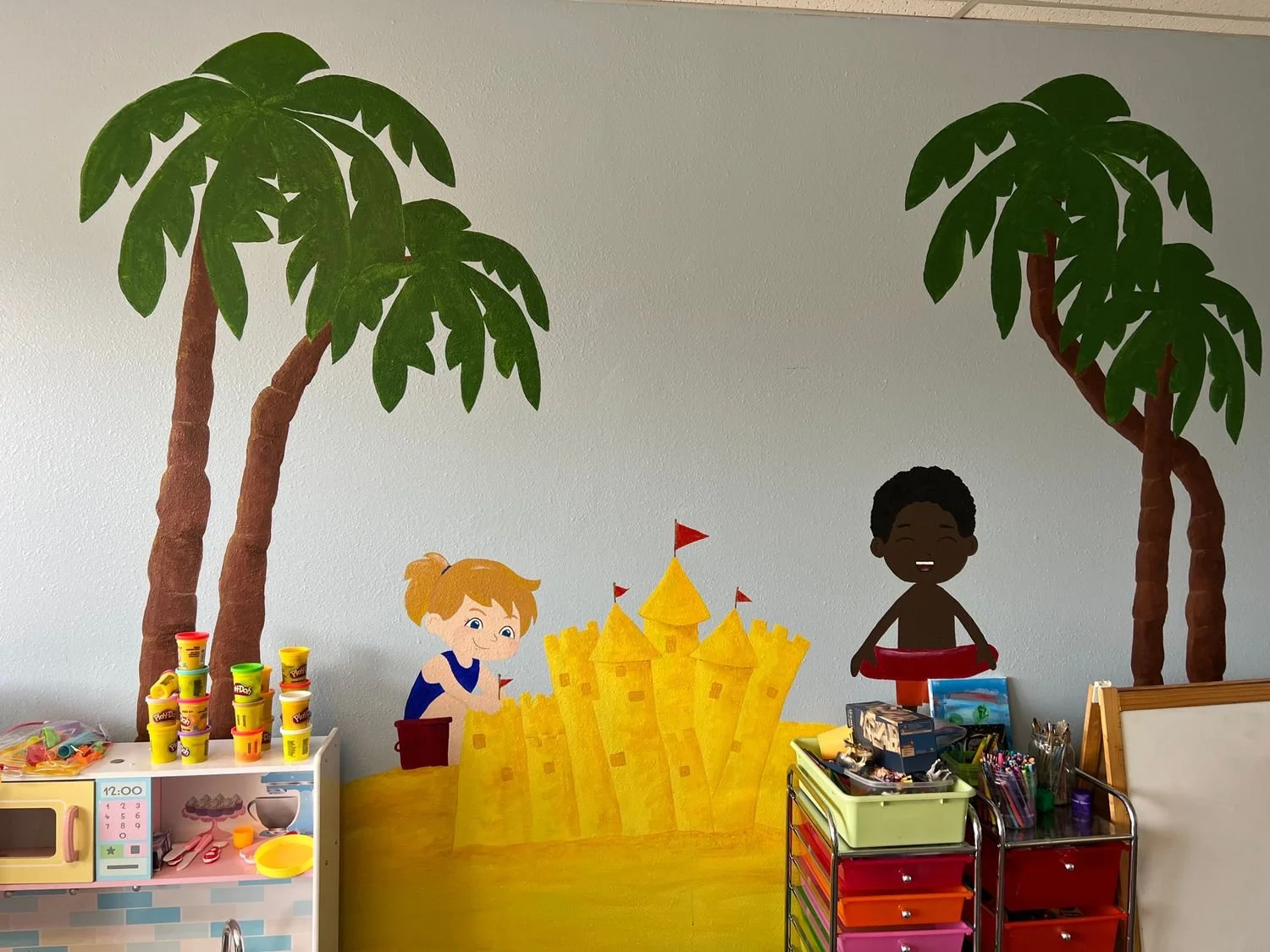 Decorative wall with painted jungle scene featuring two large trees, two children playing with a yellow sandcastle, and toy storage units.
