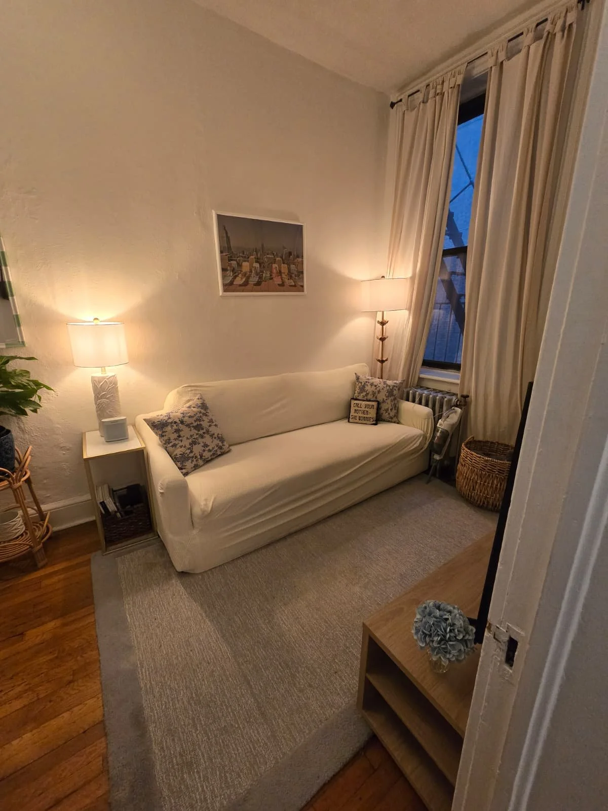 A cozy living room with a white sofa, floral pillows, two lamps, a framed cityscape picture, a wicker basket, a small side table with a potted plant, and beige curtains covering a window, with hardwood and carpeted flooring.