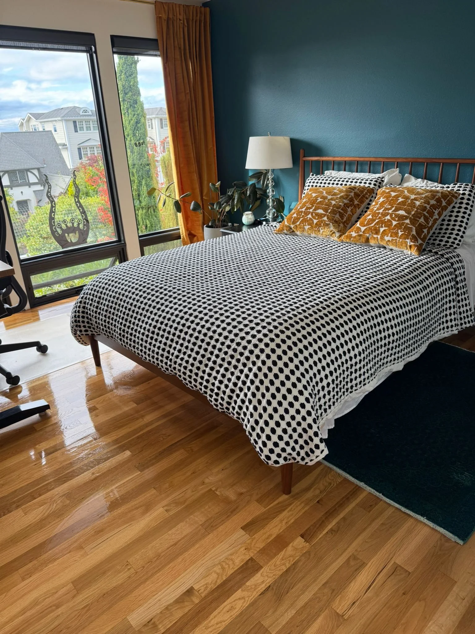 A bedroom with a bed covered in a black and white polka dot blanket, two gold patterned pillows, a teal wall, a bedside table with a lamp and plant, a window with orange curtains showing houses and trees outside, and a hardwood floor.